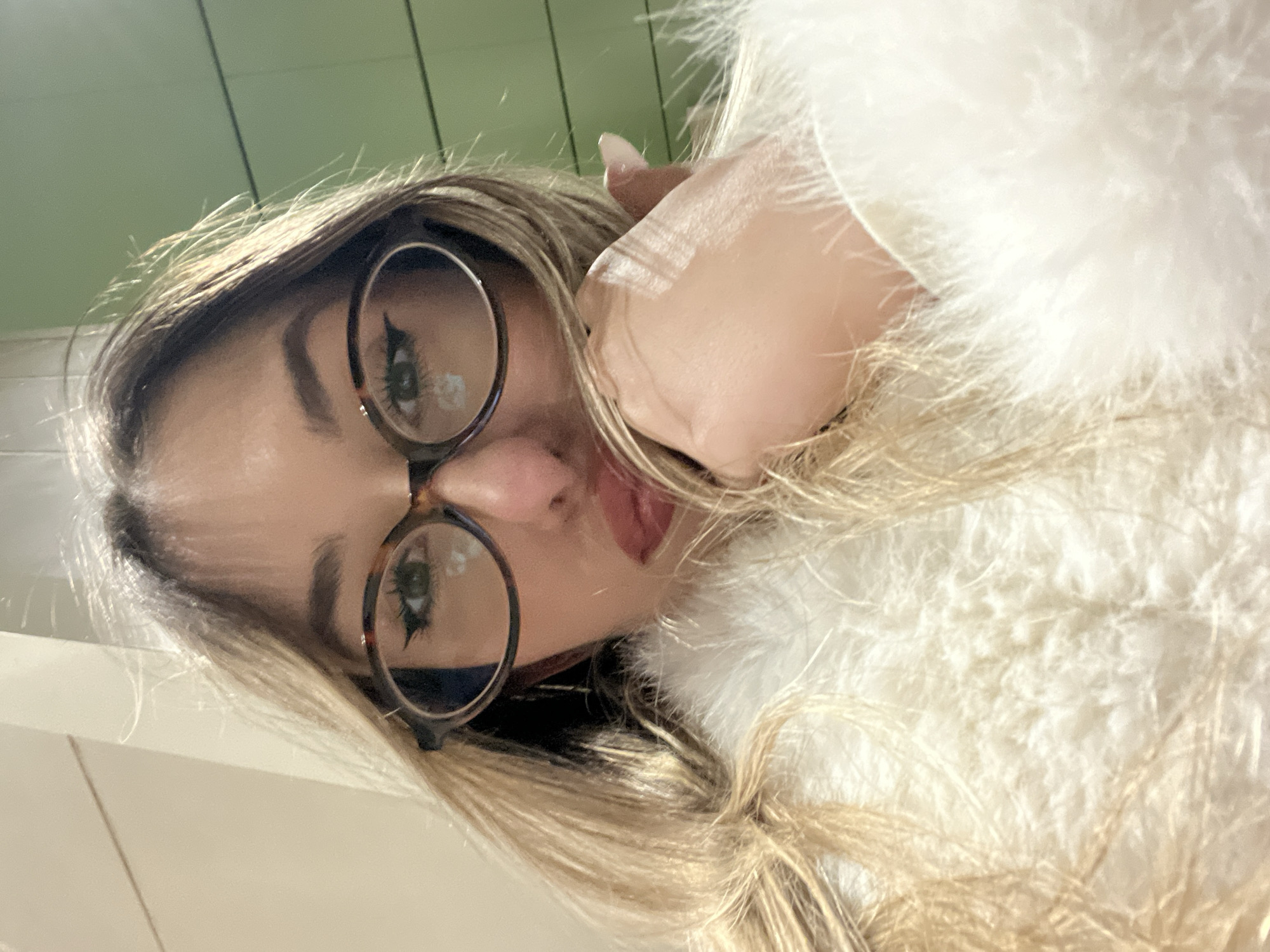 A fair-skinned person with blonde hair and round glasses is angled in a selfie shot. They are wearing a fuzzy white garment. The background appears to be a combination of white and green surfaces. The location in Sofia, Bulgaria, cannot be determined from the provided image.