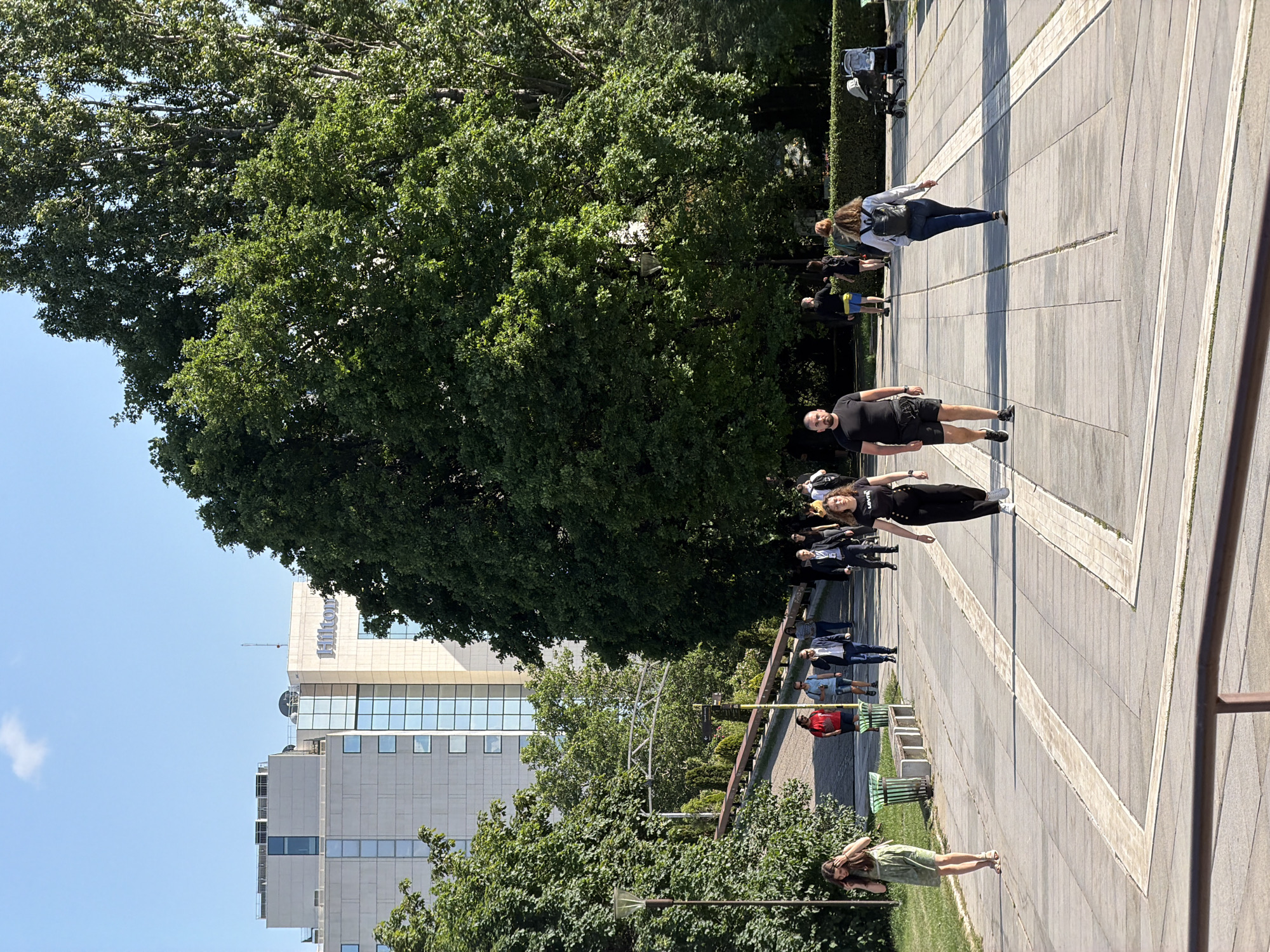 The image depicts a sunny, outdoor scene in Sofia, Bulgaria, likely near the Hilton Sofia hotel, which is visible in the background. A large tree dominates the upper portion of the frame. A paved area with geometric patterns is present, and people are walking along it. Some are individually strolling, while others appear to be in small groups. There are benches and greenery along the edge of the paved area, and some fencing or railing is also visible. A person in a green dress is standing at the edge of the scene in the bottom left of the frame.