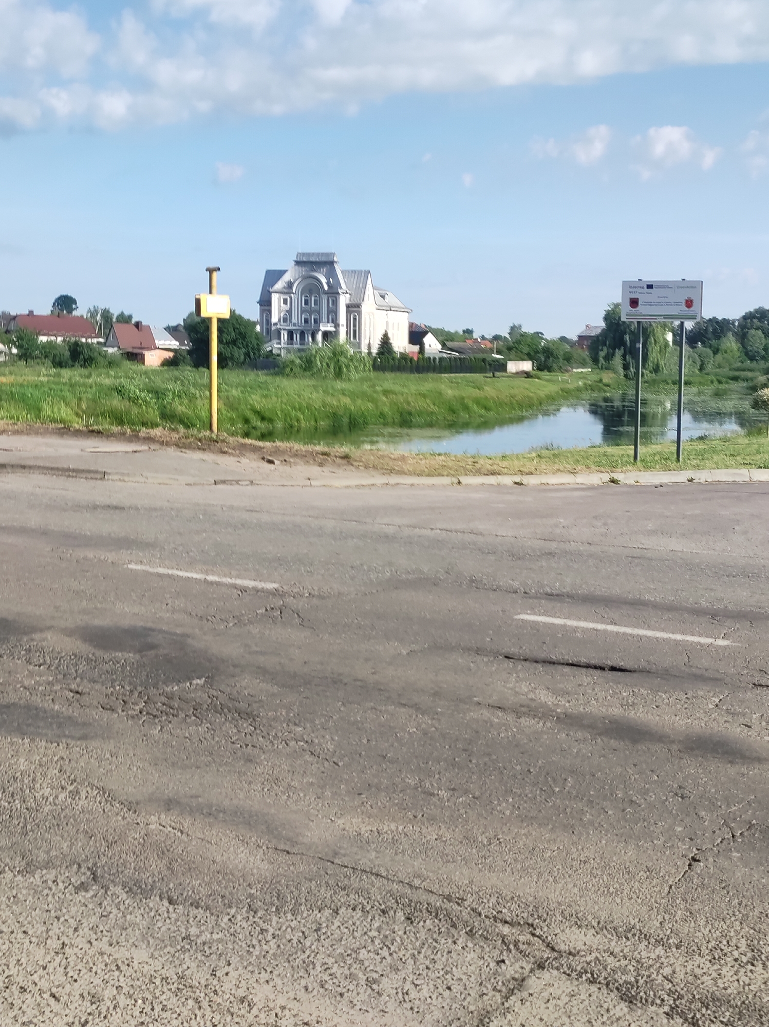 The image captures a serene daytime scene featuring a body of water reflecting the sky and surrounding greenery. In the foreground, a weathered asphalt road with visible cracks and a white lane marking stretches across the frame. To the left of the road, a yellow pole with a small, square sign stands in a grassy area. Behind this, a picturesque landscape unfolds with lush green vegetation bordering the water, which is dotted with aquatic plants.
Further back, a grand, multi-story building with a distinct architectural style, featuring arched windows and ornate detailing, dominates the horizon. To the left of this building, several residential houses with red-tiled roofs are nestled among trees. On the right side of the frame, a billboard on poles displays information, including what appear to be coat of arms and text in a European language, possibly indicating a border or local information. The sky above is a vibrant blue, adorned with scattered white clouds, suggesting fair weather. The overall impression is one of a quiet, possibly rural, setting on a bright day.