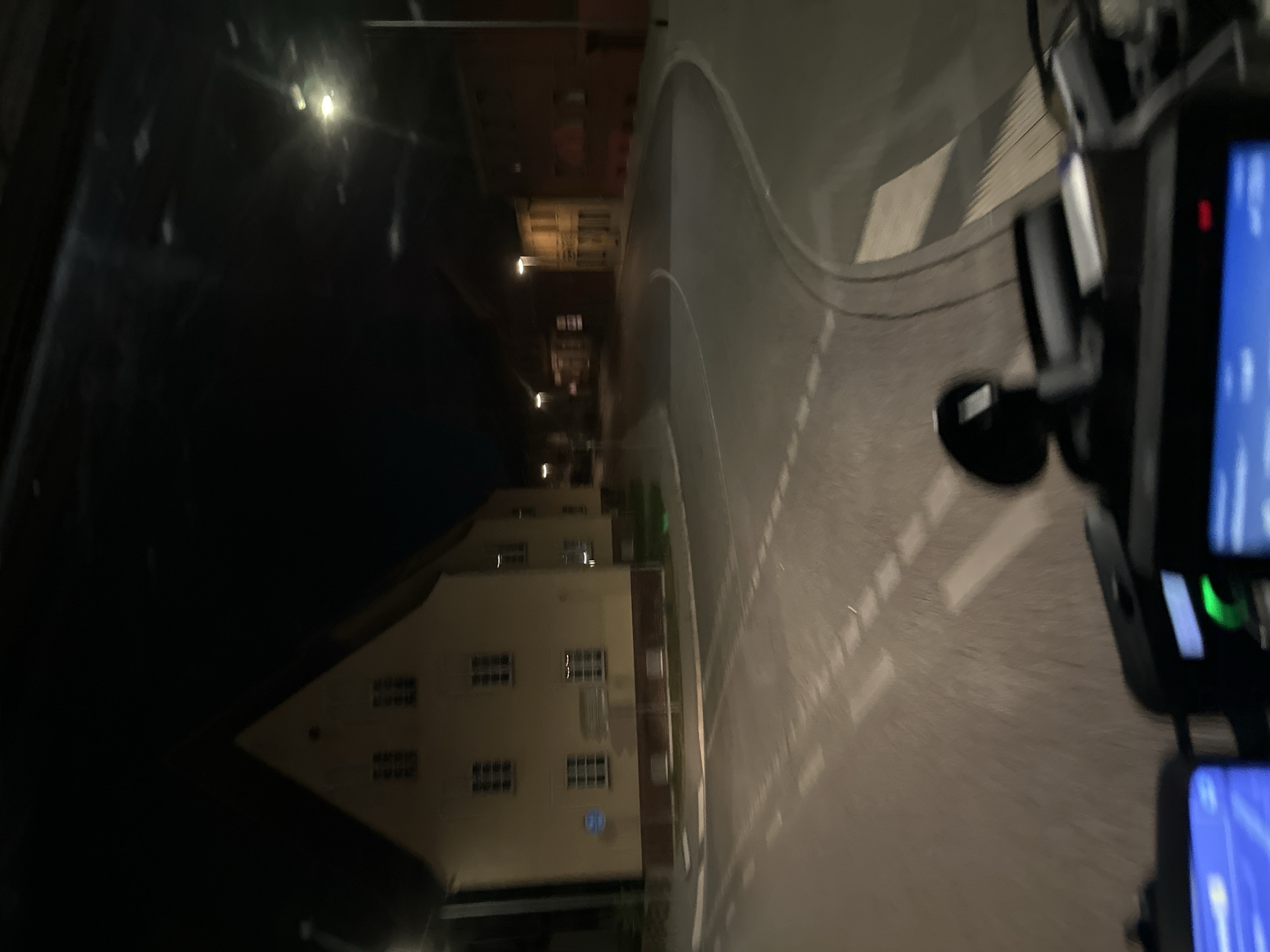 The image depicts a nighttime view from what appears to be a vehicle, possibly a motorcycle or scooter, as evidenced by the handlebars and dashboard visible in the foreground. The scene is a street in Lübben (Spreewald), Germany. On the left side of the street is a multi-story building with a distinctive gabled roof, appearing to be old architecture. The building is illuminated by streetlights, casting a warm glow on its facade, which has multiple windows. Across the street, on the right, are other buildings, one of which is brick and appears to be a commercial property or residence. Streetlights are visible throughout the scene, illuminating the road and pavement. The road itself has visible lane markings. The overall impression is of a quiet, perhaps historic, part of a town at night.