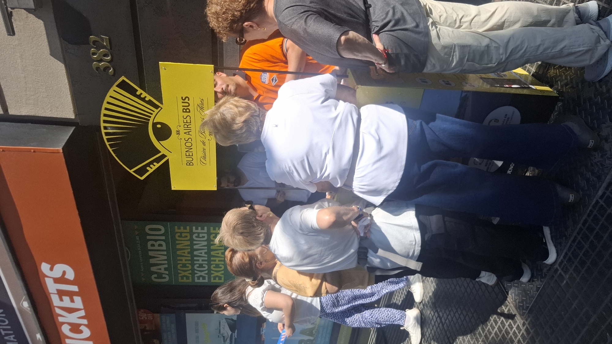 The image shows a Buenos Aires city scene. The location is evidenced by a yellow sign reading "Buenos Aires Bus, Clásico de Buenos Aires." A sign indicates a cambio (exchange) service. Other signs advertise "Tickets" and a "50% Off" promotion. Several individuals are visible in the scene, some standing and others partially obscured by others. The building displays the number "382". The environment suggests a tourist area or bus stop, likely in central Buenos Aires.