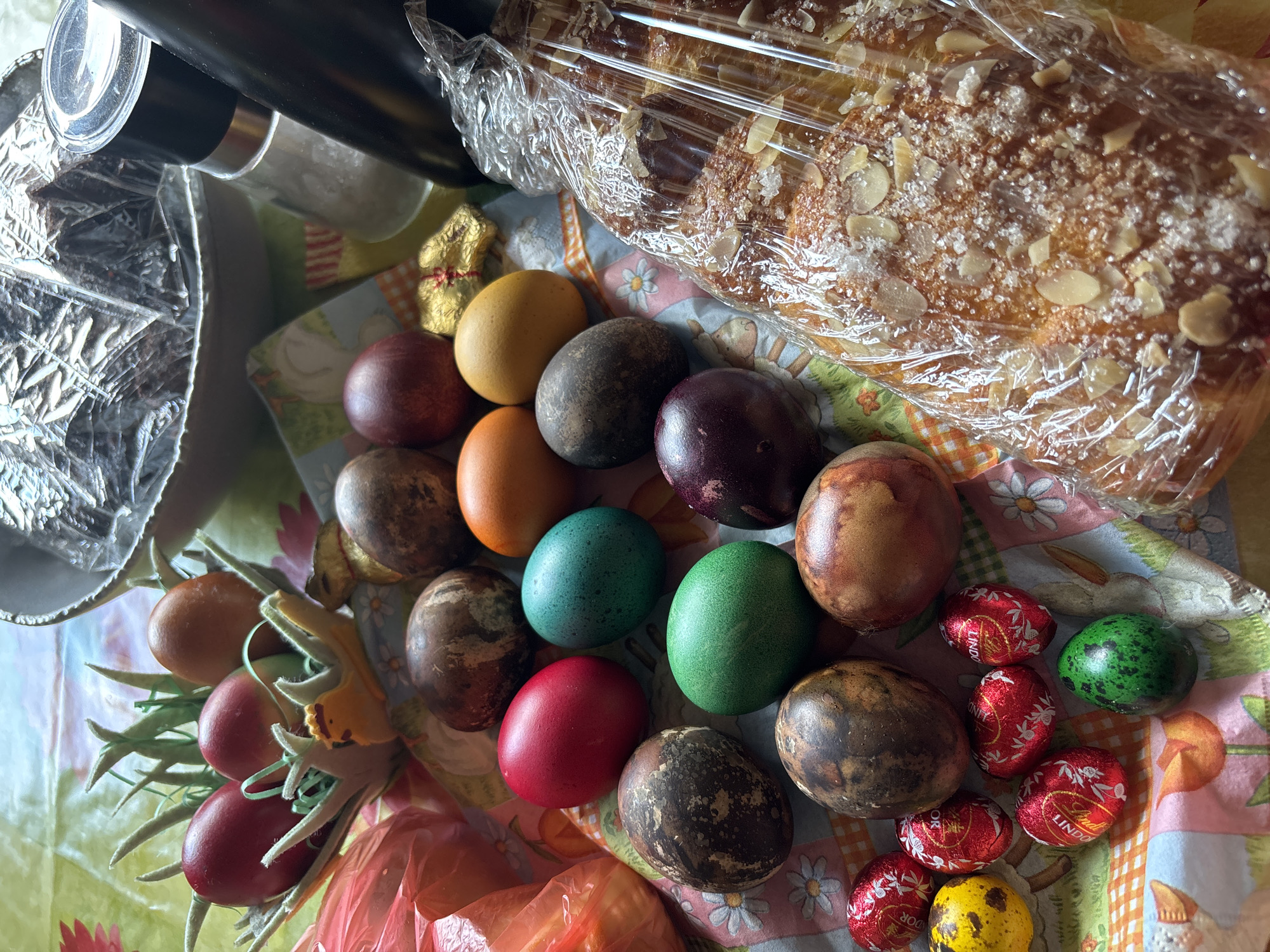 A top-down shot captures an Easter feast laid out on a patterned tablecloth featuring daisies, rabbits, and carrots. Dominating the scene is a wrapped sweet bread, its surface covered with almonds and sugar. Adjacent to the bread is a collection of dyed Easter eggs in various colors: yellow, orange, brown, red, blue, and green. Some eggs exhibit a mottled or stained appearance. Additionally, there are several small, foil-wrapped chocolate eggs, primarily red with "Lindor" branding, and one yellow egg with dark speckles. To the left, a cluster of red and orange eggs is arranged with artificial green foliage, suggesting a decorative arrangement. A portion of a silver and black object, possibly a coffee carafe, is visible in the upper left corner. The setting and content suggest a traditional Easter celebration, though the location in Goritsa, Bulgaria is not discernible from the visual information provided. There are no people visible in the scene.
