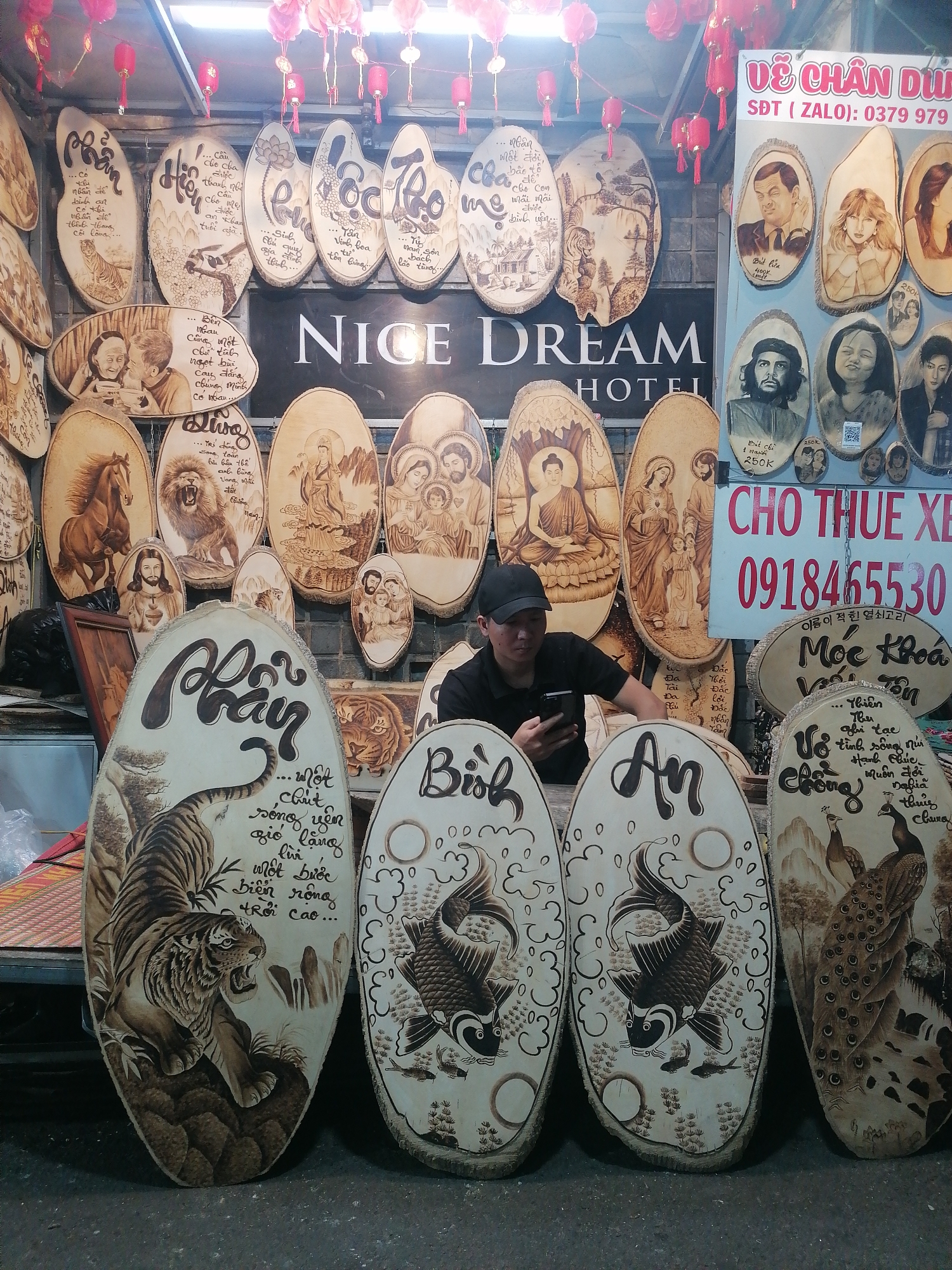 A man stands in front of a display of wood-burned artwork in what appears to be a street market in Da Lat, Vietnam. He is wearing a black shirt and a black baseball cap, looking at his mobile phone. The artwork consists of a variety of oval wooden plaques, each with a different image burned into the surface. Some plaques feature animals like tigers, lions, and peacocks. Other plaques depict people, including portraits and scenes of religious figures. The plaques are arranged on shelves and the ground around the man. Above the artwork, there's a sign that reads "NICE DREAM HOTEL." A poster with portraits for sale has phone numbers written on it. Lanterns decorate the top of the scene.