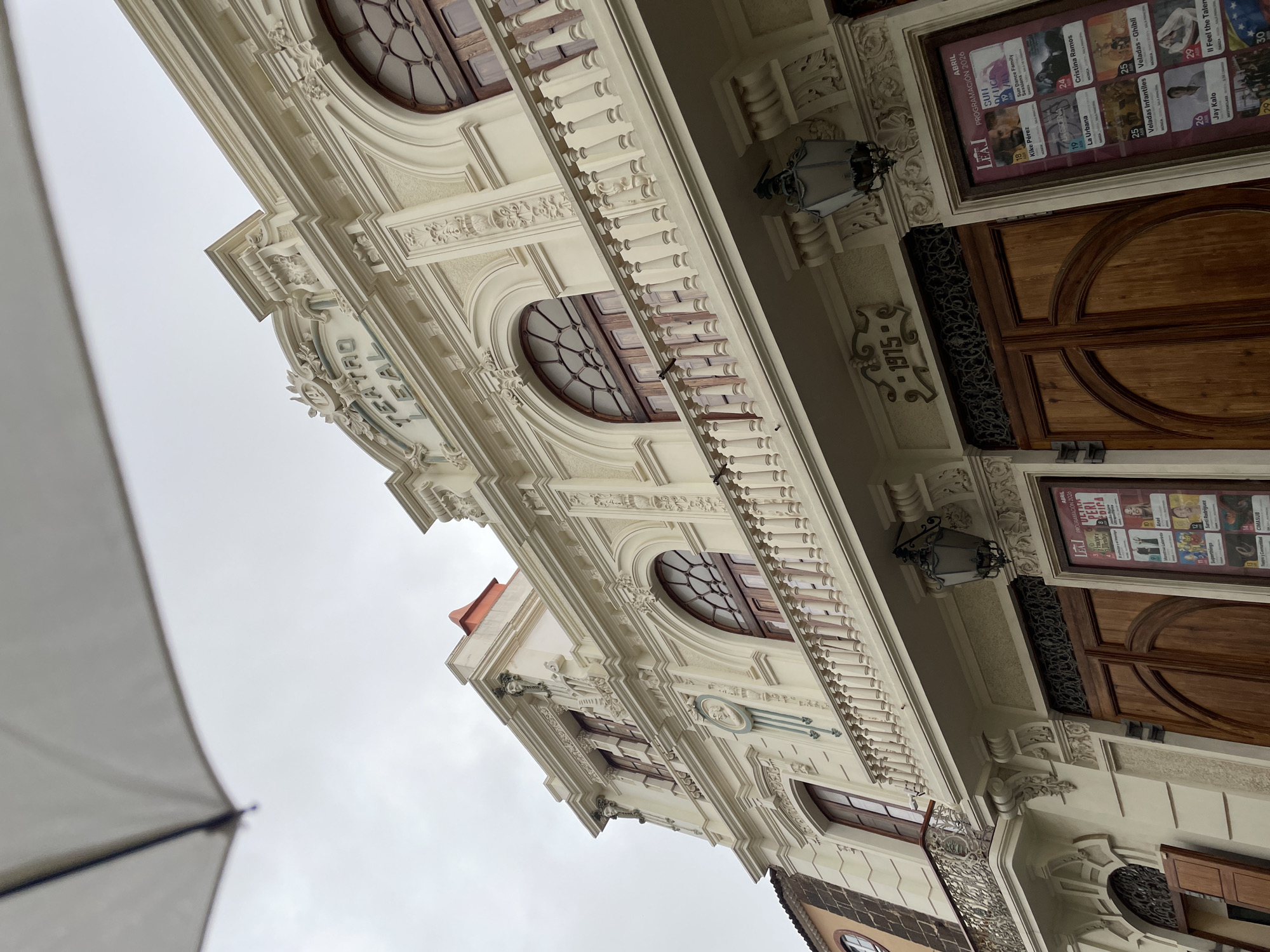 An upward-angled, slightly rotated view captures the ornate facade of the Teatro Leal in La Cuesta, Spain, under an overcast sky. The grand building is cream-colored and features classical architectural elements, including decorative carvings, balustrades, and rounded windows with intricate wooden grilles. Two large, dark wooden doors are visible on the right, flanked by elegant lanterns.
Visible text "TEATRO LEAL" is prominently displayed on the building's facade, confirming its identity. The year "1915" is carved into the stonework, likely indicating its construction date. Advertising posters for upcoming events are affixed near the entrance. These posters display "ABRIL PROGRAMACIÓN 2023" and "ABRIL PROGRAMACIÓN 2026," listing various performances such as "Superthings," "Santi Rodriguez," "Kike Pérez," "Sun Dance Family Sessions," "La Urbana," "Cristina Ramos," "Veladas Infantiles," "Veladas - Ghibli," "Jay Kalo," "II Feel the Talent," and "MASTERCLASS."
A large, white or light-colored umbrella is partially visible in the bottom-left foreground, suggesting the photographer might be sheltered from rain or sun. The scene is quiet, with no people actively engaging in events, but the posters indicate cultural activities take place there. The grey, cloudy sky suggests daytime, possibly a cool or damp day.