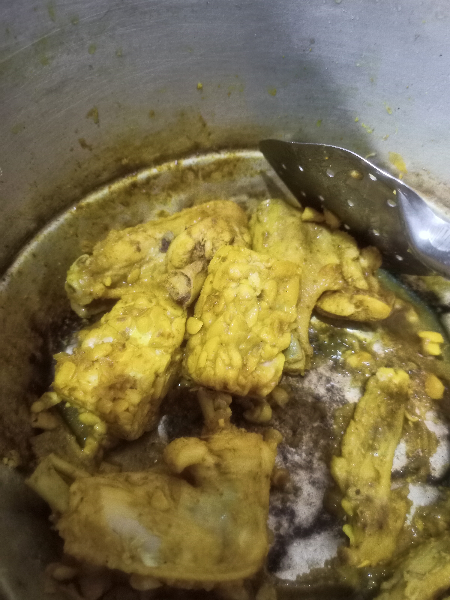 A close-up, overhead shot shows cooked chicken pieces with a yellow spice coating in a pot. The chicken appears to be in a stew or curry, with some liquid and scattered spices visible. A slotted metal utensil rests on the right side of the pot, partially submerged in the food. The pot itself is metallic with some discoloration and residue on the interior. The image is likely from a kitchen in Karangtalun, Indonesia, where such dishes are commonly prepared.