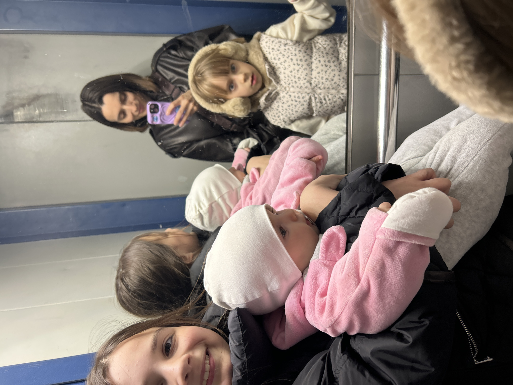 A rotated image shows a group of people, primarily females, with two children. The setting appears to be a train, based on the metal bar and the interior. The image is taken in front of a mirrored surface reflecting the scene.
The foreground features a smiling girl, a baby wearing a white hat and pink clothing, and a person's hand. The mirror reflects a woman holding a phone, another child with a fur-trimmed hood, and other details of their clothing and the train interior. There is no verifiable information to indicate that the image was taken in Moscow, Russia.