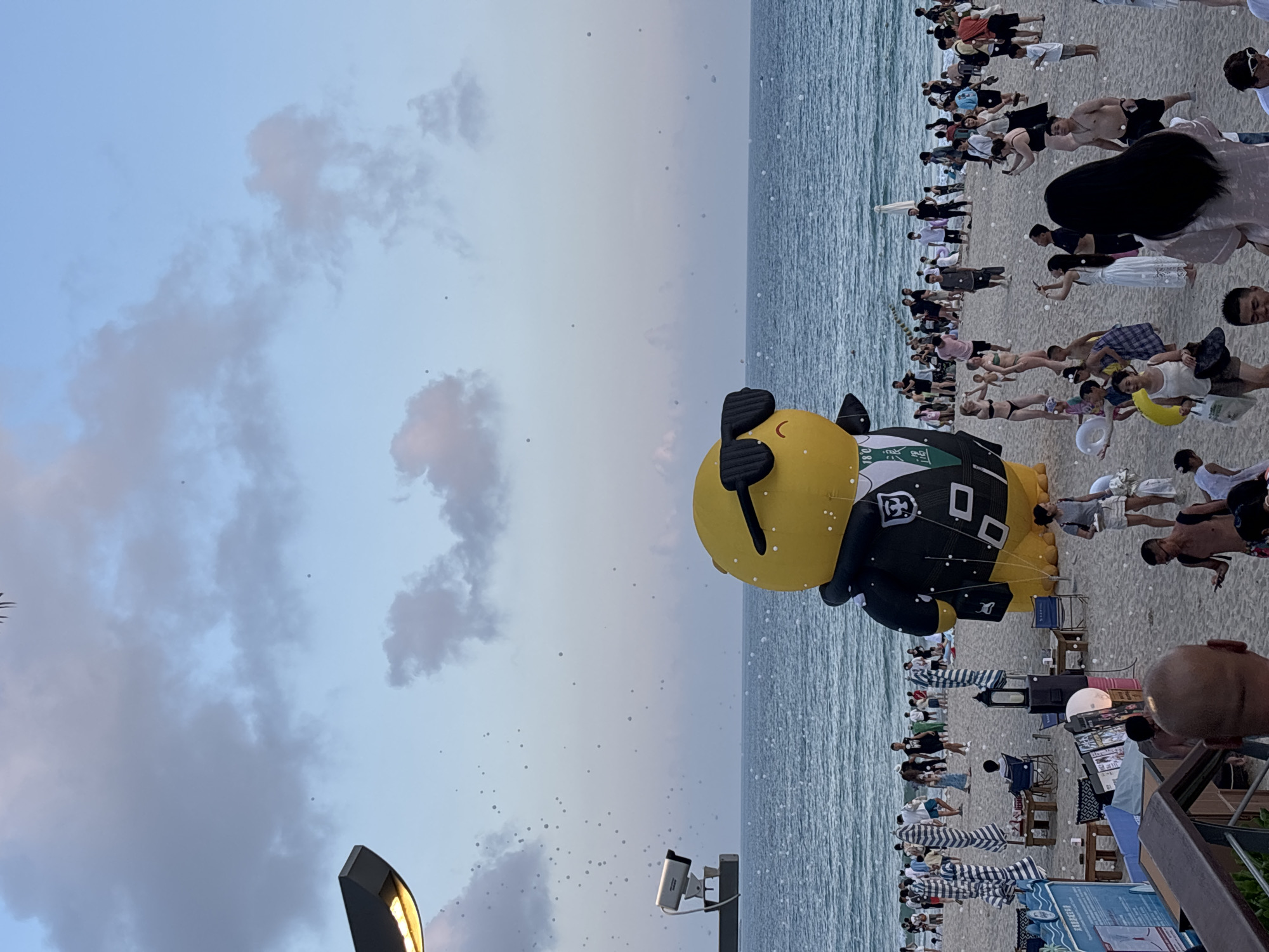 A scene unfolds on a sandy beach, likely in the city of Jiyang District, China, under a twilight sky with scattered clouds. In the foreground, a large, inflatable yellow figure sporting dark sunglasses and a black jacket with white trim stands prominently. The beach is populated with numerous people, some walking, some standing, and some interacting. A calm sea stretches out behind the beach, with gentle waves lapping at the shore. Beach umbrellas and seating areas are visible, suggesting a leisure or event setting. A lamppost with a light fixture and a security camera are positioned on the left side of the frame, overlooking the scene. Small, white, spherical objects are scattered across the sand and in the air, potentially indicating confetti or bubbles. The overall atmosphere appears festive and relaxed.