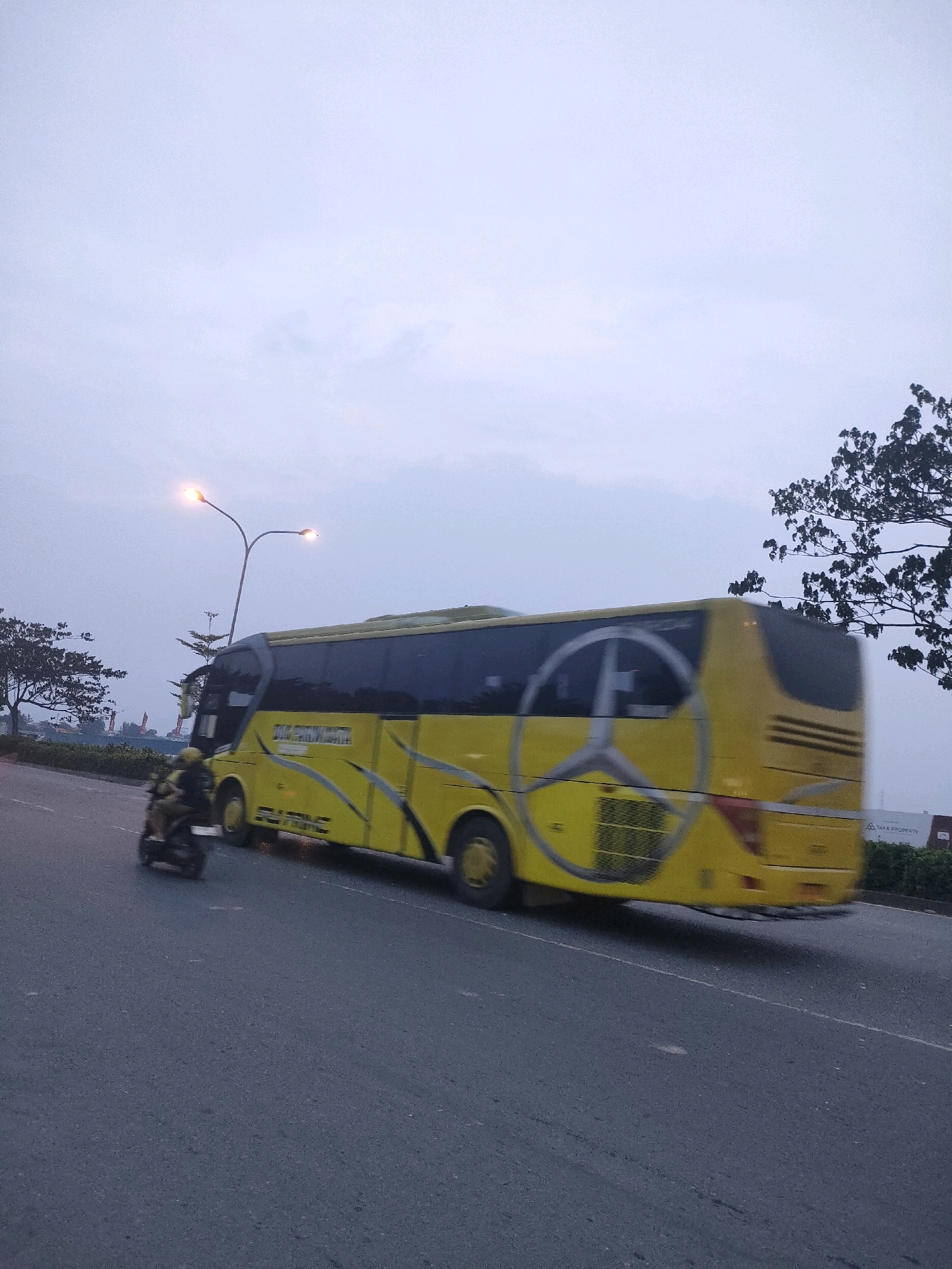 A bright yellow bus with a large silver Mercedes-Benz emblem on its side is captured in motion on a paved road. The bus has black tinted windows and appears to be a passenger bus, with the text "PUTRIMANI" visible in yellow lettering along its side. In front of the bus, a motorcyclist wearing a helmet is also in motion. The scene takes place in Batam, Indonesia, under an overcast sky. Streetlights with two lamps are visible in the background, indicating it may be dawn or dusk. Trees and some distant buildings are also present in the environment, suggesting an urban or suburban setting. The overall impression is one of a transportation hub or a busy road in Batam.