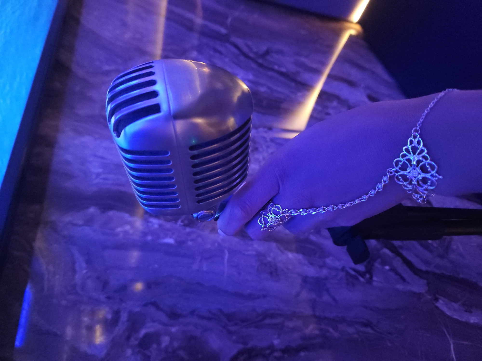 A hand adorned with an ornate silver hand chain, holding a silver microphone in a purple and blue lit room. The wall is marble. The location is Unknown, Unknown.