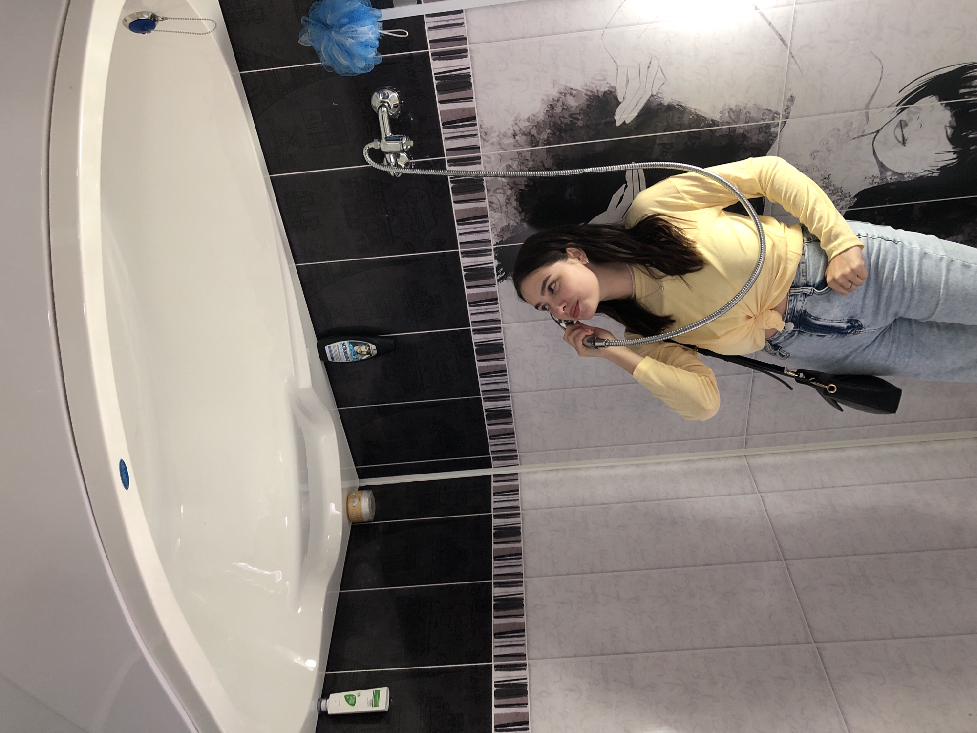 A young woman with dark hair, wearing a yellow long-sleeved shirt tied at the waist and light-wash jeans, stands in a shower stall. She is holding a shower head to her ear as if on a phone and looking upwards. The shower stall has black tiled walls with a decorative strip of grey and white tiles above the white, patterned wall tiles. A shower faucet and hose are visible. On the black tiles, a blue pouf hangs to the right of the faucet, and two bottles of toiletries are mounted to the left. The woman has a black purse slung over her shoulder. This scene is not identifiable as being located in Izhevsk, Russia.