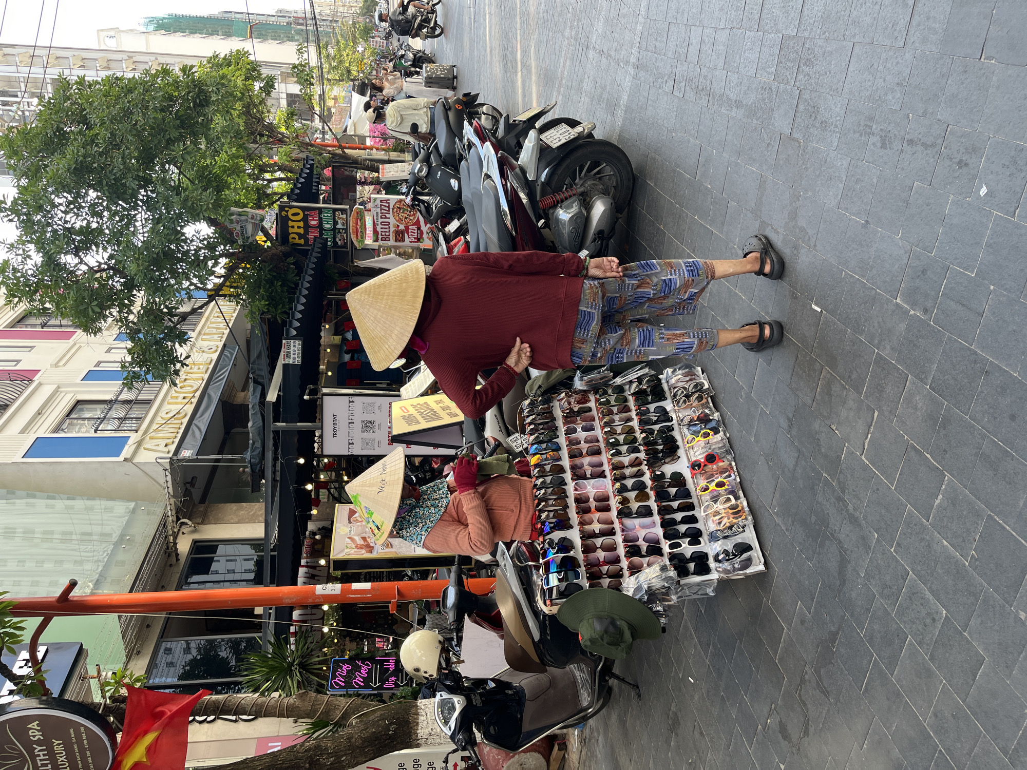 The scene takes place on a sidewalk in the city of Đà Nẵng, Vietnam. Two vendors wearing traditional Vietnamese conical hats are selling sunglasses from tables. One vendor stands wearing a red long-sleeved shirt and patterned shorts, while the other is seated, wearing pink gloves. Numerous motorcycles are parked nearby along the sidewalk. In the background, there are buildings with shops and restaurants with various signs. One sign advertises "BELLO PIZZA". A Vietnamese flag is visible. A sign for "Healthy Spa Luxury" is partially visible on a pole in the foreground.