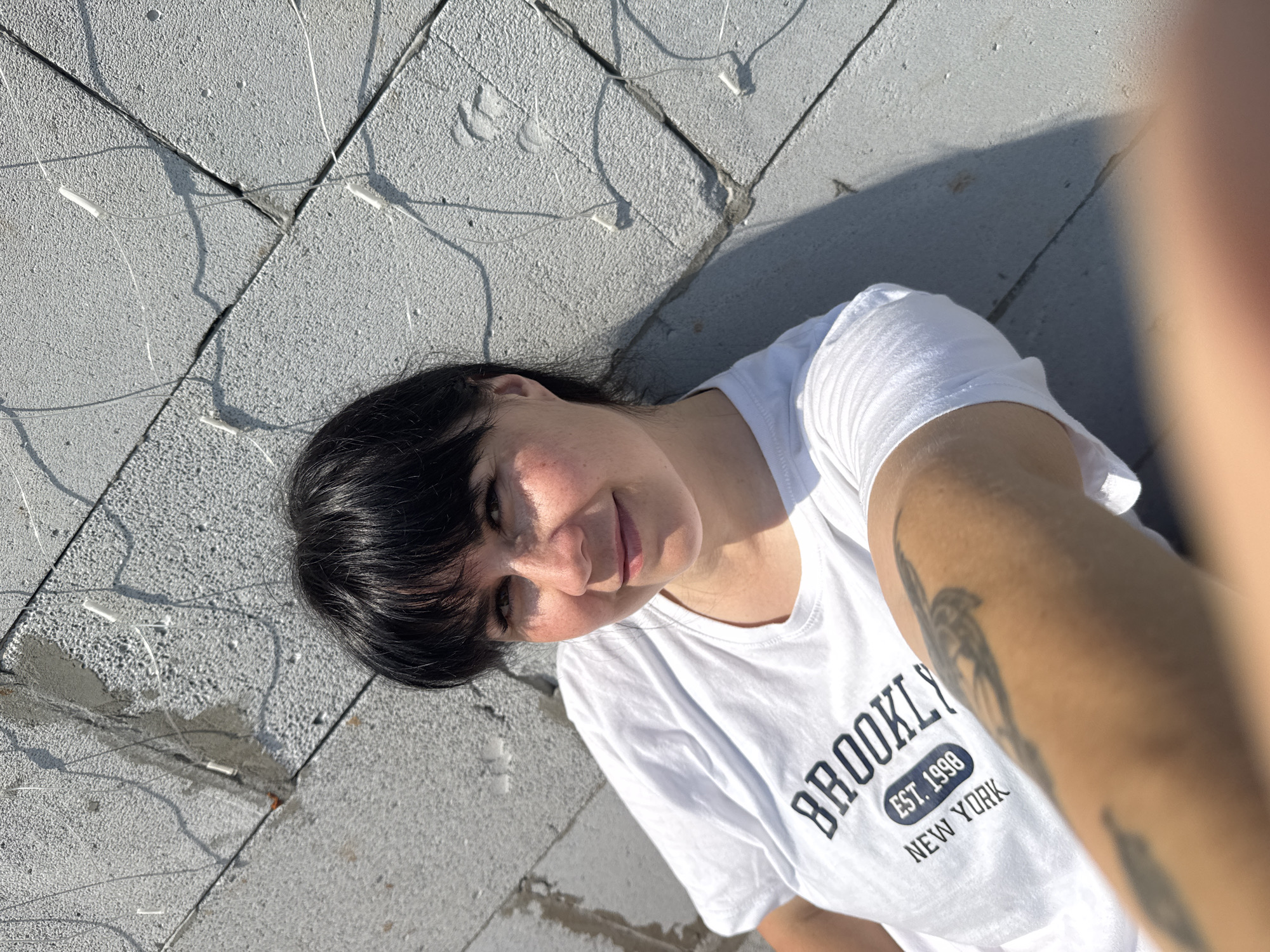 In Zav'yalovo, Russia, a woman with dark hair and bangs is captured in a selfie. She is wearing a white t-shirt that reads "BROOKLYN EST. 1998 NEW YORK." A tattoo is visible on her left arm. The woman is positioned against a rough, grey concrete wall, which appears to be part of a building under construction, as indicated by visible wires and sealant. The scene is illuminated by bright sunlight, casting shadows on the wall.