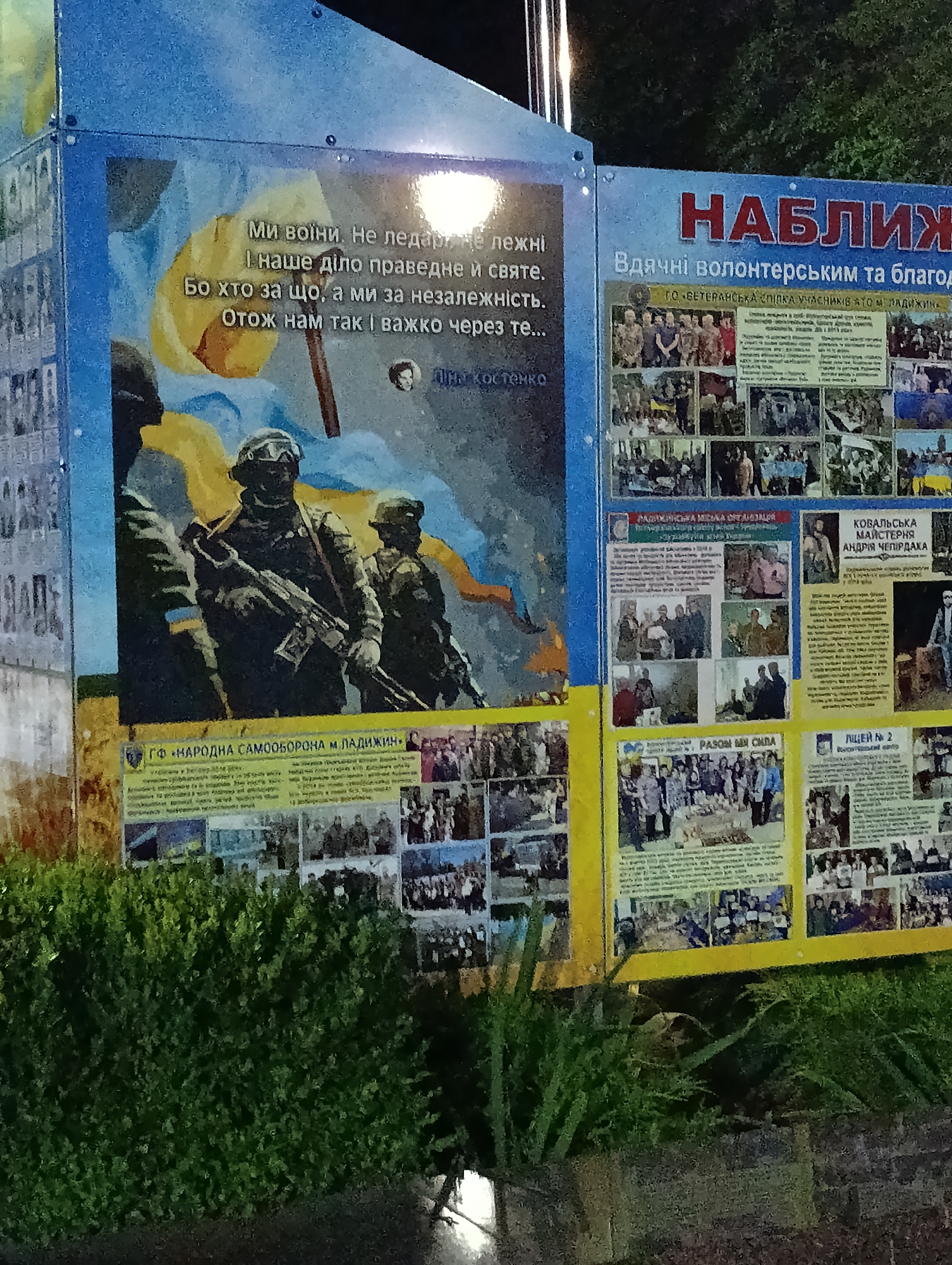 This is a photograph of an outdoor memorial display in the city of Ladyzhyn, Ukraine. The display consists of large panels with text and images, likely commemorating individuals and events related to the conflict.
The leftmost panel features a painted mural of three soldiers in military attire, one in the foreground holding a rifle. Behind them, a Ukrainian flag motif is visible. Text in Ukrainian is superimposed on the mural.
The central and right panels are collages of photographs and text. The central panel displays a large headline in red: "НАБЛИЖЕНИЕ" (NABLYZHENIE, meaning "Approach" or "Closeness"). Below this, in smaller text, it reads "Вдячні волонтерським та благодійним організаціям" (Grateful to volunteer and charitable organizations). This panel contains numerous smaller photographs of people, groups of people, and possibly events.
The rightmost panel also contains multiple photographs and Ukrainian text. A headline in red reads "НАГОРОДЖЕННЯ" (NAGORODZHENNYA, meaning "Awards" or "Decorations"). This section appears to be dedicated to recognizing individuals or groups. Another section of the right panel has a headline that reads "ЛИЦЕЙ №2" (LYCEI №2, likely referring to Lyceum No. 2).
The display is situated outdoors, with lush green bushes and foliage in the foreground obscuring the base of the panels. The background appears to be dark, suggesting the photograph was taken at night or in low light conditions. The panels are mounted vertically, likely on a structure. The overall impression is a public tribute or information display.
