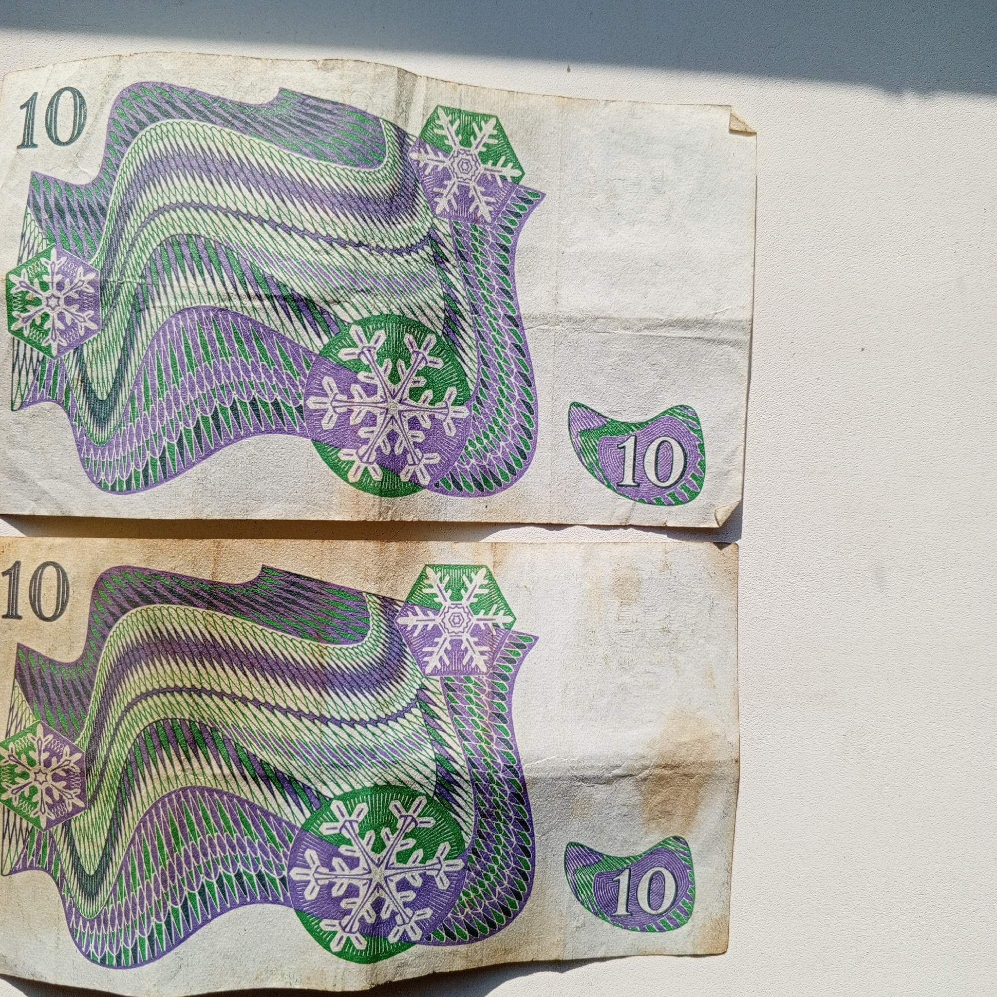 The image displays two crumpled ten-unit currency notes, likely banknotes, lying flat on a textured, off-white surface. The notes are positioned one above the other, with a visible gap between them. Both notes feature intricate purple and green patterns, resembling waves or stylized feathers, and a snowflake-like emblem. The numeral "10" is prominently displayed on each note in a dark gray or black color, both on the upper left corner and within a smaller, curved design element on the right side. The lighting suggests a strong light source casting shadows, with a dark shadow falling from the upper left of the frame onto the surface and the top note. The surface itself appears to be a wall or a similar textured material. The image does not contain any people, actions, or interactions. The location of Sayanogorsk, Russia, is not depicted or implied in the image.