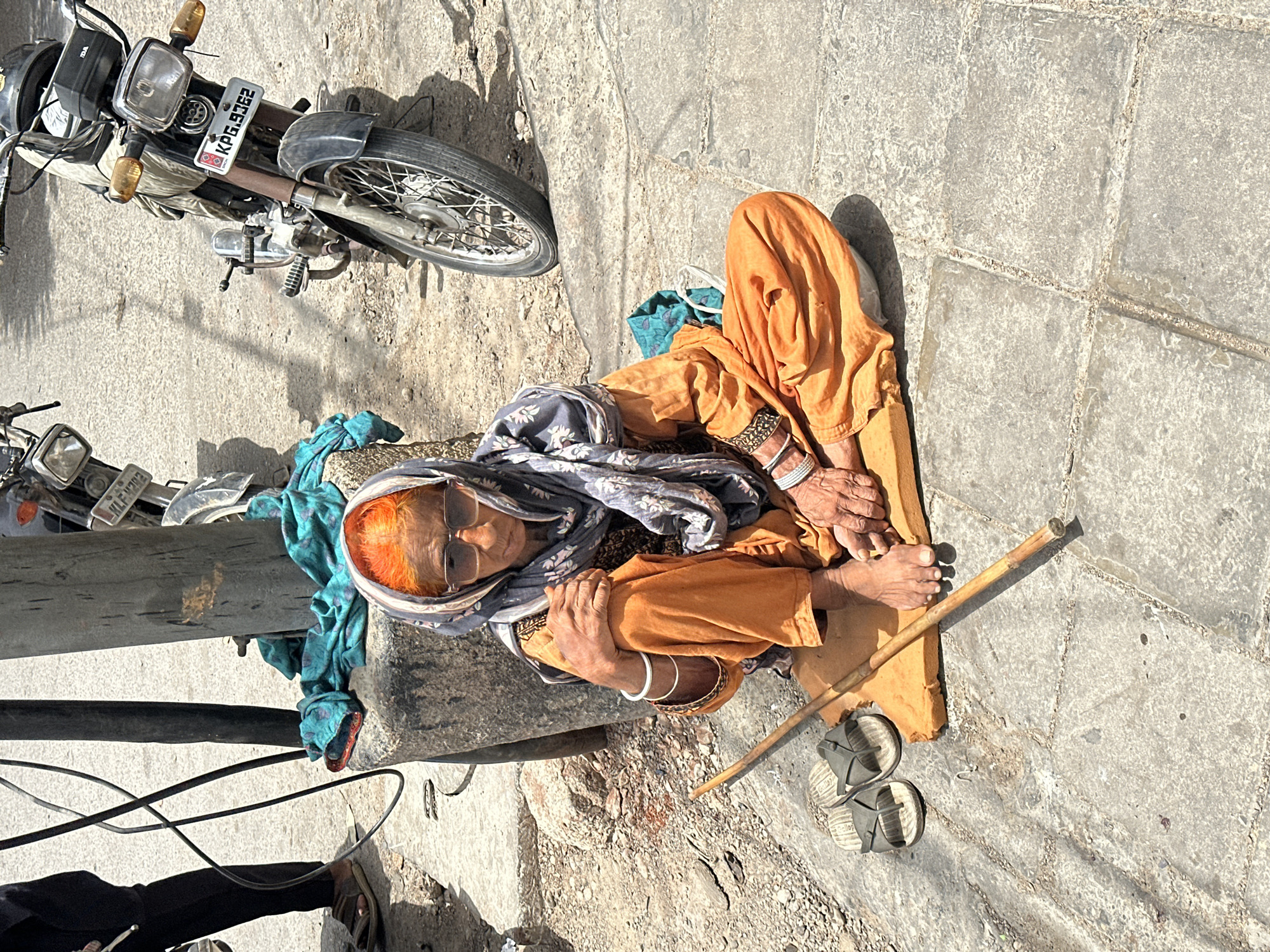 An elderly woman with bright orange hair sits on a thin yellow mat on a tiled sidewalk in Karachi, Pakistan. She is wearing an orange salwar kameez and a grey patterned scarf. Her arms are crossed over her knees, and she has several bangles on her wrists. She wears large sunglasses. A thin wooden stick lies in front of her on the ground, parallel to her legs. To her left are a pair of dark grey sandals. Behind her, resting against a concrete pillar, is a pile of folded teal and white fabric. A motorcycle is parked to her right, its front wheel visible, and its license plate reads "KPG.9362". In the background, a black wire hangs from a pole. A person's legs and feet, wearing dark trousers and brown sandals, are visible on the left side of the frame.