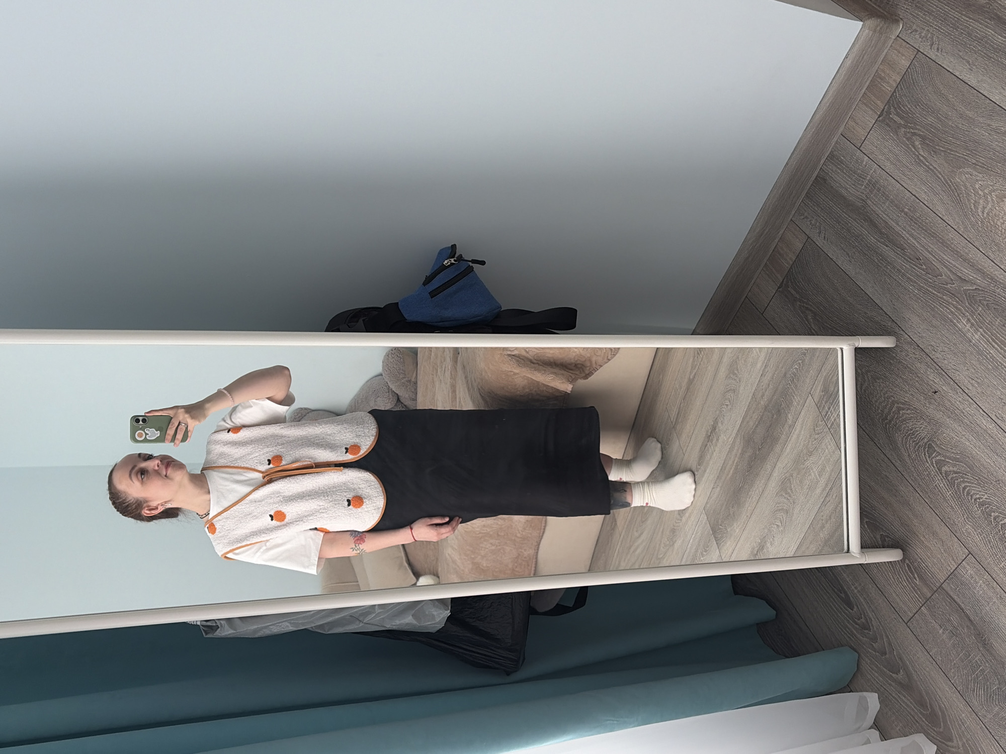 A mirror selfie of a woman standing in a room. The woman, with hair pulled back and fair skin, is wearing a white t-shirt, a black jumper dress, and white socks. She is holding a cell phone in her right hand, taking the picture.
Behind her, a couch with beige pillows is visible. Above the mirror, a blue backpack sits on a shelf against a light blue wall. The flooring to the right of the mirror is a wood-patterned laminate. Below the mirror, partially obscured, are blue and white curtains. The location is presumed to be Прикамский, Russia based on implied information.