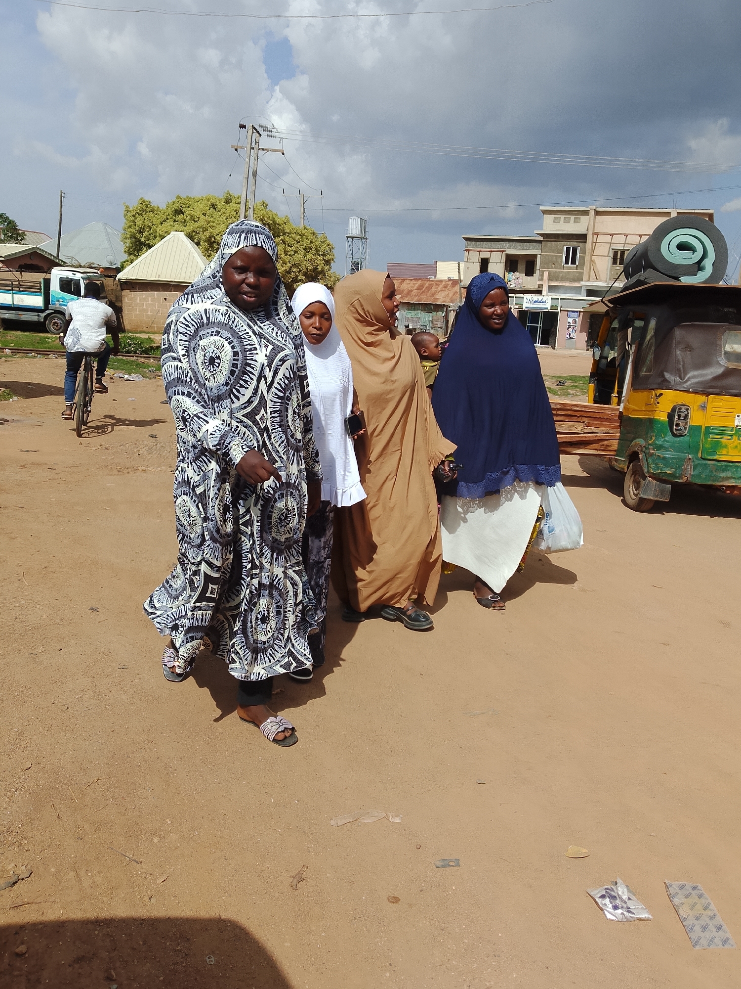 In an outdoor urban setting, four individuals are walking on a dirt road under a partly cloudy sky. The foreground features the lower bodies of three women. The woman on the left wears a long, patterned dress and beaded sandals. Next to her, a woman in a white hijab and dress is partially visible. To their right, another woman is dressed in a tan hijab and long dress, holding a white bag. Behind her, a woman in a dark blue hijab and white skirt is also visible. In the background, a person rides a bicycle, a white truck is parked, and several buildings and a three-wheeled vehicle are present. The ground is covered in dirt and scattered debris.