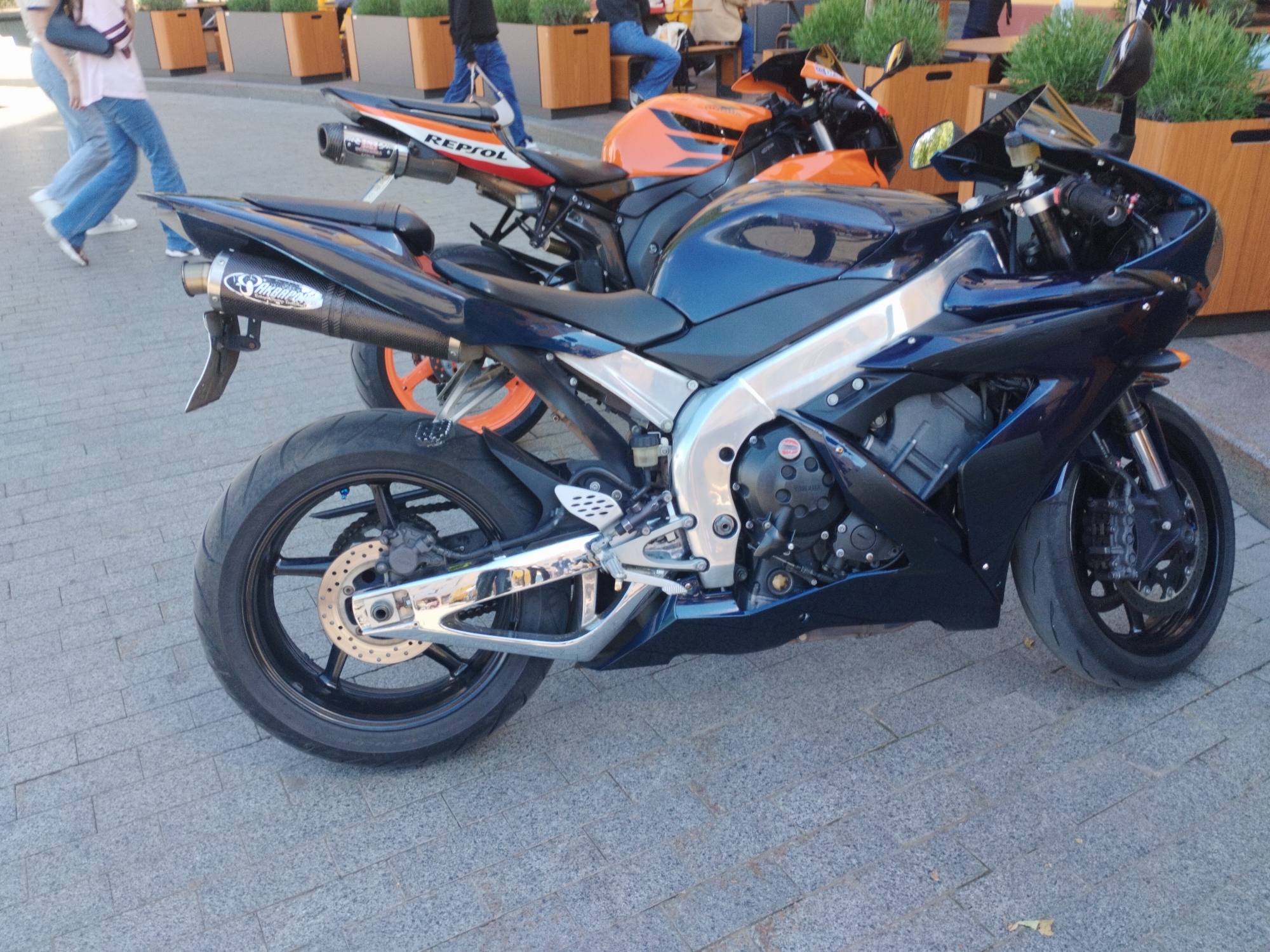The image depicts two parked motorcycles on a paved surface. The foreground motorcycle is dark blue, while the one behind it is orange and white with "Repsol" branding. Pedestrians are visible in the background near wooden planters with green plants. The location is identified as being in Odesa, Ukraine.