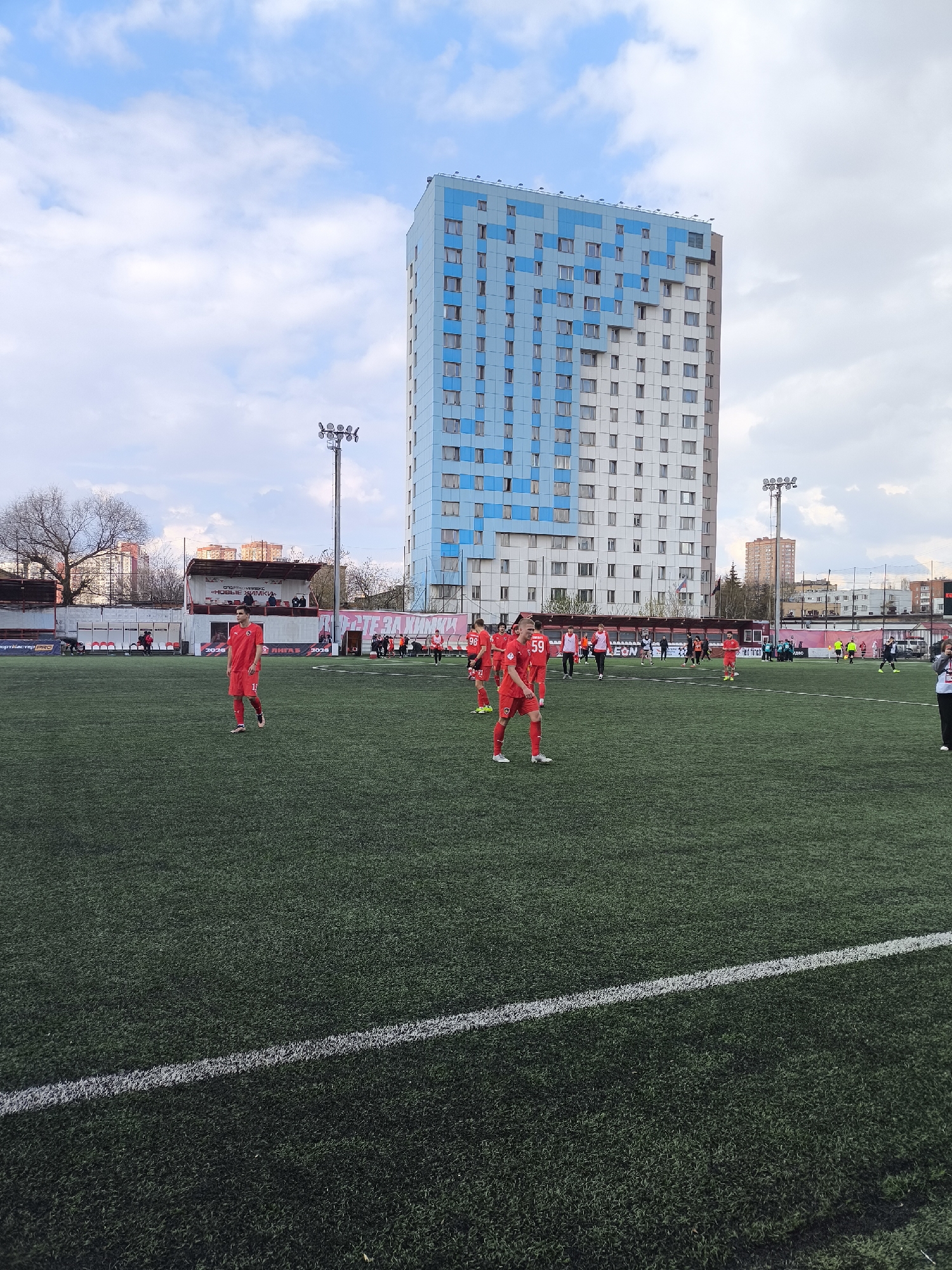 The image shows a soccer field in Khimki, Russia. The field is green artificial turf with white boundary lines. Several soccer players in red uniforms are scattered across the field. In the background is a large, blue and white apartment building. There is a small stadium bleacher area visible behind a fence. Light poles are placed on the side of the soccer field and in the background. The sky is partly cloudy.