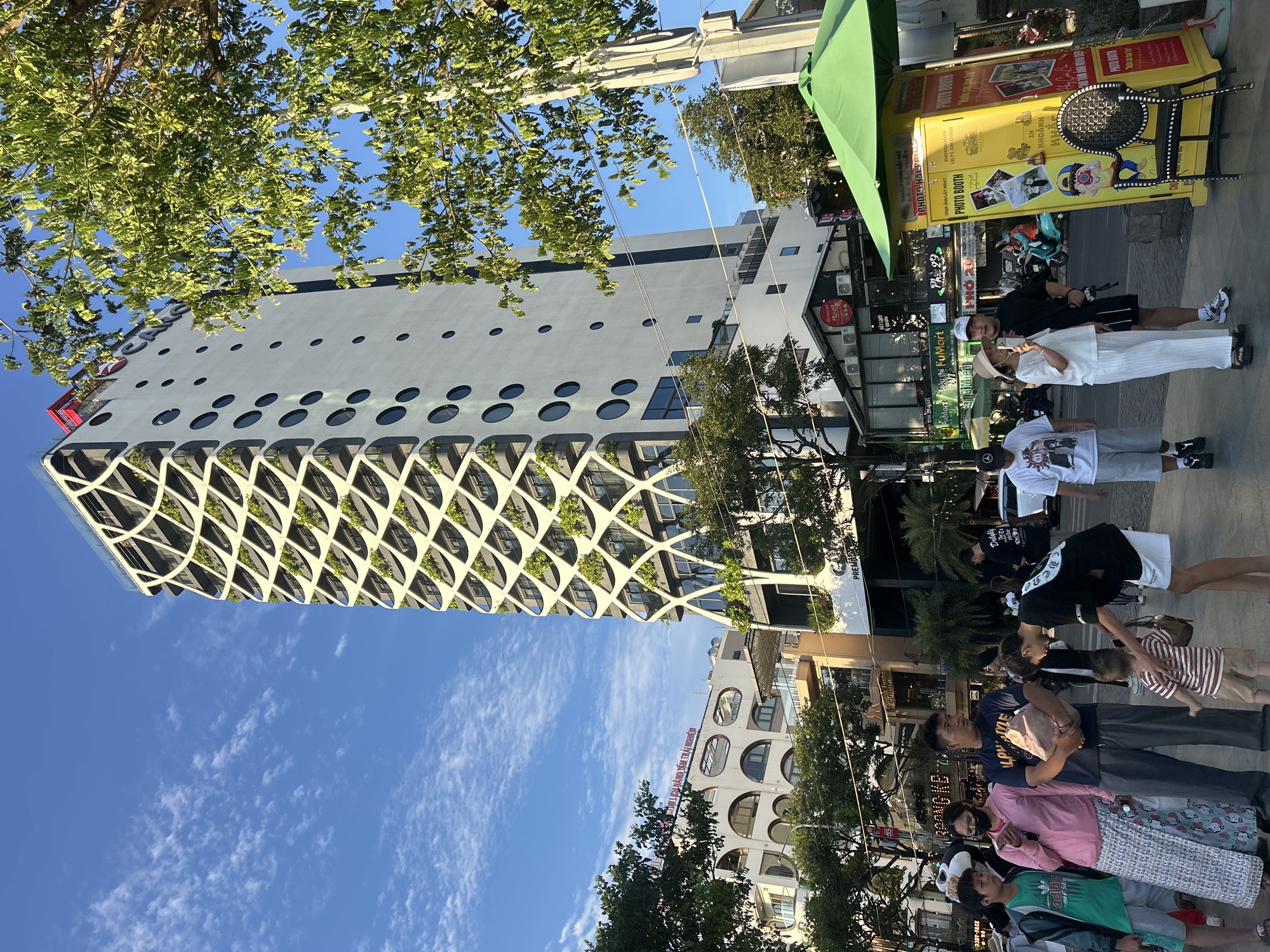 The image depicts a bustling street scene in Đà Nẵng, Vietnam, specifically featuring an apartment building of the Premier Village Danang Resort located at 99 Võ Nguyên Giáp, Ngũ Hành Sơn District. The scene is illuminated by clear daylight under a blue sky with scattered white clouds.
In the background, a tall, multi-story white building with a distinctive facade dominates the frame. Its left side features a latticework of curved white elements creating balcony spaces, many of which contain vibrant green plants. The right side of the building is smooth and white, punctuated by numerous circular windows of varying sizes arranged in vertical and horizontal alignments. A red sign at the building's pinnacle displays the text "PREMIER VILLAGE DA NANG". To the left of this prominent structure, a lower, lighter-colored building with multiple arched windows and balconies is partially visible, bearing a sign in Vietnamese that reads "KHU DU LỊCH NẰNG TÂM TRẢI NGHIỆM".
At street level, a group of approximately ten individuals, including adults and at least two children, are present on the sidewalk. On the left side of the foreground, a young boy in a green Adidas t-shirt and grey shorts stands next to a woman wearing a pink long-sleeved top, a patterned long skirt, and a black face mask, who is looking at her phone. Beside them, a man in a dark blue t-shirt and grey pants holds a light brown bag. Further to the right, a man in a black t-shirt and black shorts, wearing a white baseball cap, bends down to interact with a small child in a striped t-shirt and light brown shorts. A woman in a white wide-leg pants outfit and a straw hat looks at her phone, and another man in a white graphic t-shirt and grey shorts walks away.
Commercial establishments line the street, with visible signs for "FUMart 24/7" and "Phở 29". A bright yellow photo booth with a green umbrella canopy stands on the right sidewalk, displaying "PHOTO BOOTH" in English and Vietnamese, along with various graphics and a black metal chair with an ornate backrest. Green-leafed trees partially obscure parts of the buildings and sky. Utility poles and overhead wires are also visible, traversing the scene.