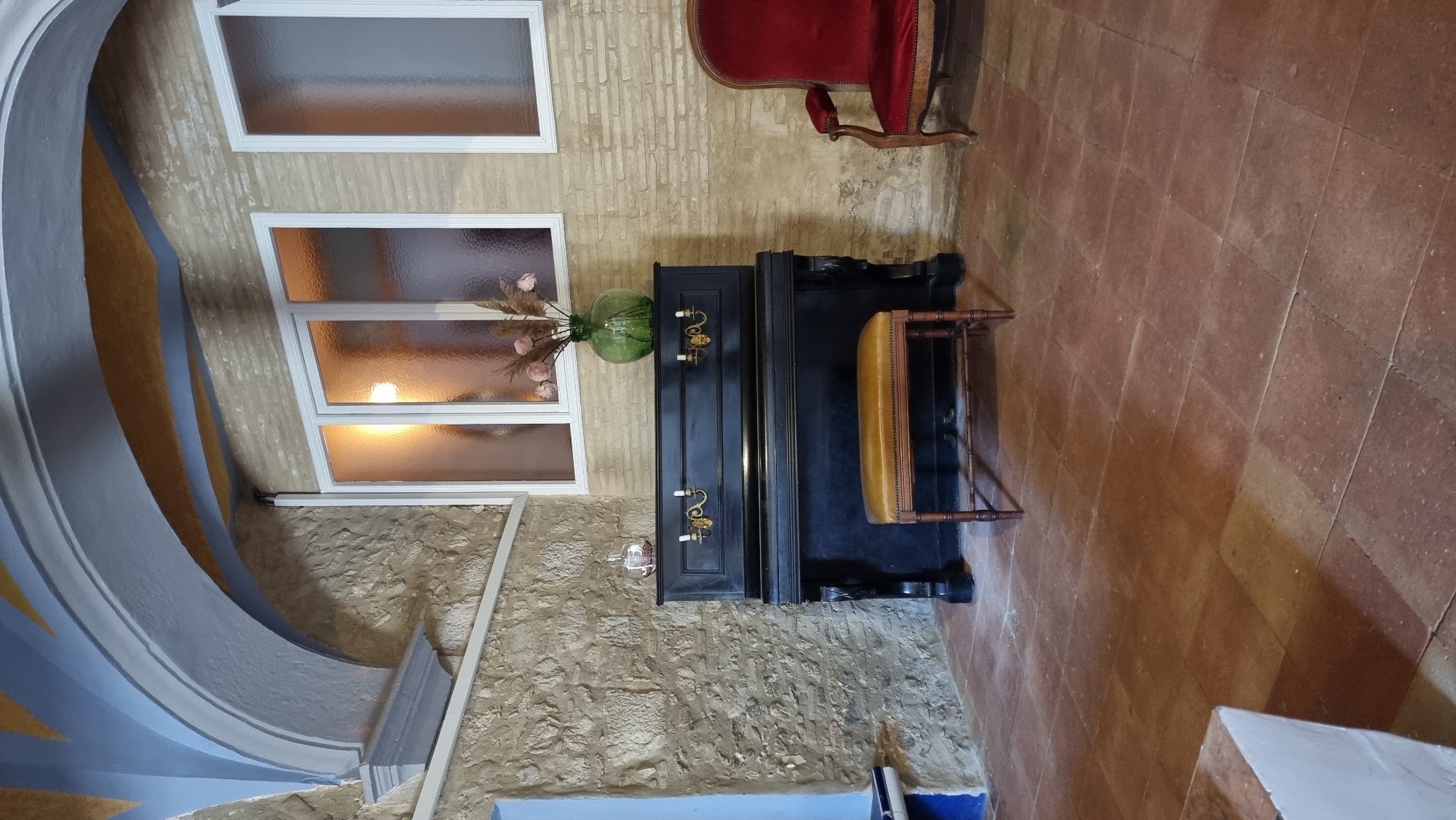 The image depicts an interior scene of a room in Castelló d'Empúries, Spain, featuring a black upright piano with a yellow upholstered bench in front of it. The piano has decorative brass candle holders on its top. To the right of the piano, a plush red armchair is visible. The room has textured stone walls and a tiled floor. Above the piano, a set of frosted windows allows light to enter, and decorative architectural elements, possibly part of a ceiling or archway, are visible in the upper portion of the image. A green vase with dried flowers sits on top of the piano. The overall ambiance is one of an antique or historic interior.