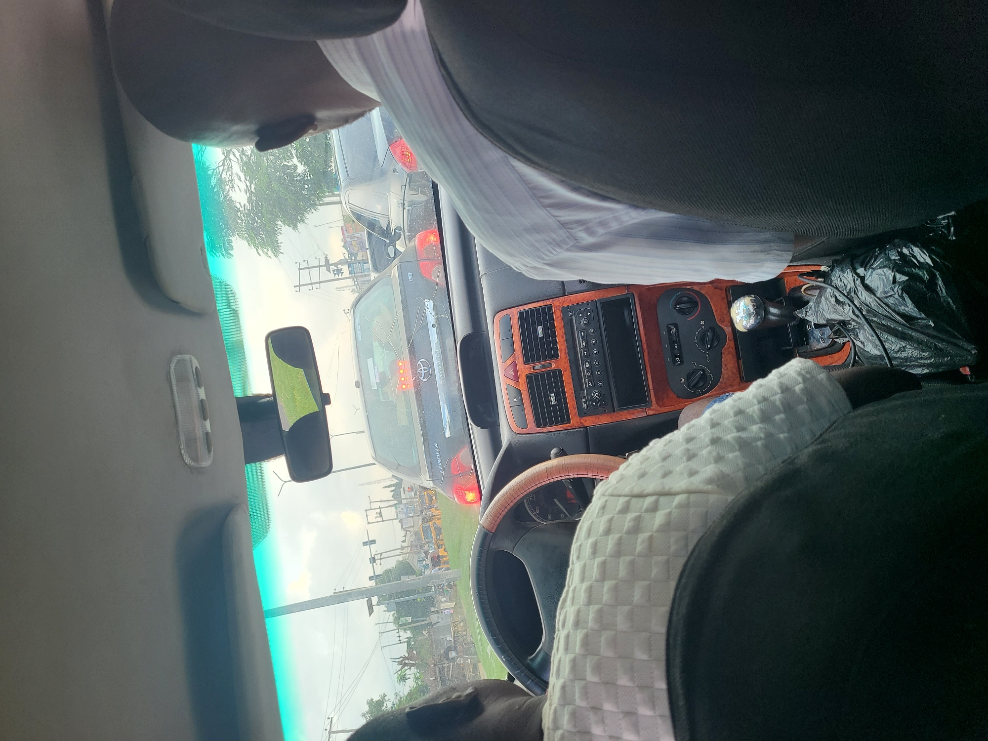 The image is a first-person perspective from inside a car in traffic in the city of Karu, Nigeria. The driver, an adult male with dark skin and a shaved head, is wearing a white patterned shirt and is visible from the back. To the right, another adult male with dark skin, wearing a light blue and white striped shirt, is also visible from the back. The car's dashboard features orange trim, a stereo system, air conditioning controls, and a gear shift. A black plastic bag is placed next to the gear shift. Outside the car, traffic is heavy, with several vehicles including a black Toyota Corolla and other cars and motorcycles visible. The sky is overcast, and there are trees and utility poles in the background. The overall impression is one of a typical urban traffic scene in Nigeria.