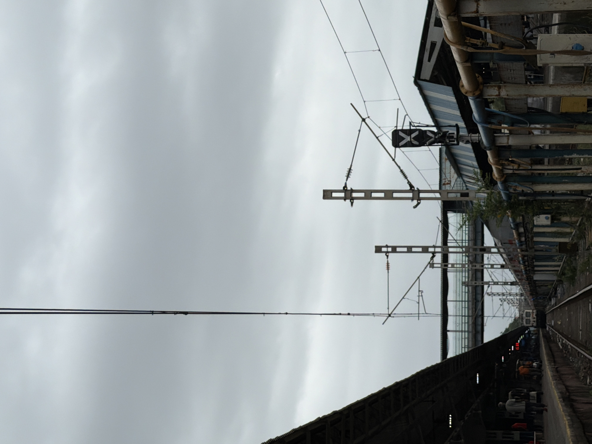The image depicts a cloudy day at a Mumbai railway station. In the foreground, railway tracks are visible, with a platform to the left and a fence to the right. The platform features a covered walkway with people walking along it. Beyond the tracks, another platform with a building and overhead pedestrian bridge can be seen. Overhead wires and signal poles are prominent throughout the scene. A train is partially visible in the distance, approaching the station. The overall environment suggests a busy, operational railway hub.