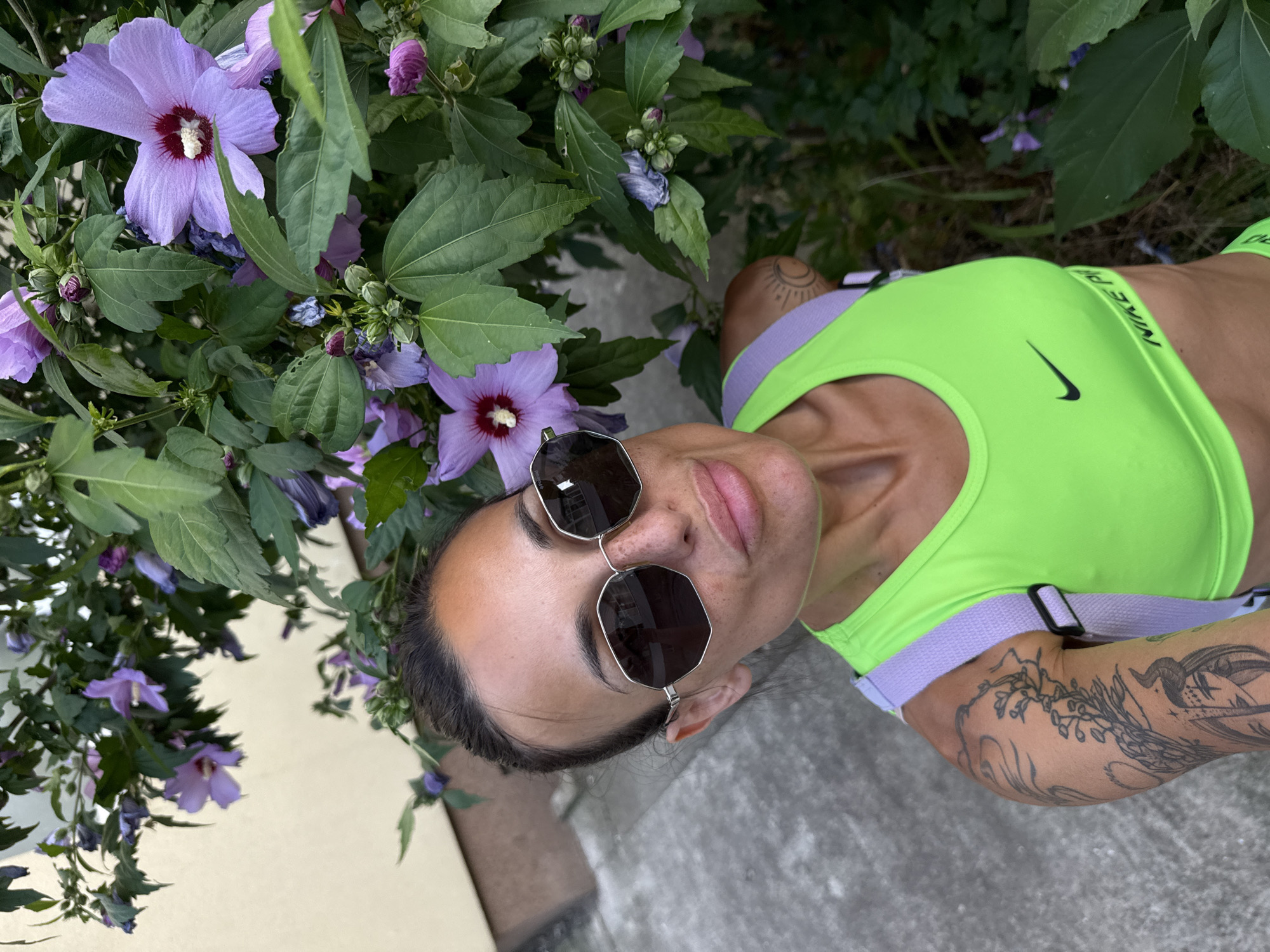 The image features a person in the foreground, lying on their back on a concrete surface. They are wearing a neon green sports bra with purple straps and have dark hair, bangs, and sunglasses. The person has tattoos on their arm and shoulder, and their face is turned towards the camera. In the background, there are purple hibiscus flowers and green foliage, suggesting an outdoor setting. The lighting and shadows indicate it is daytime, possibly late afternoon. The overall mood is relaxed and casual. There is text visible on the sports bra that reads "NIKE PRO". The image appears to be taken in Pleven, Bulgaria.