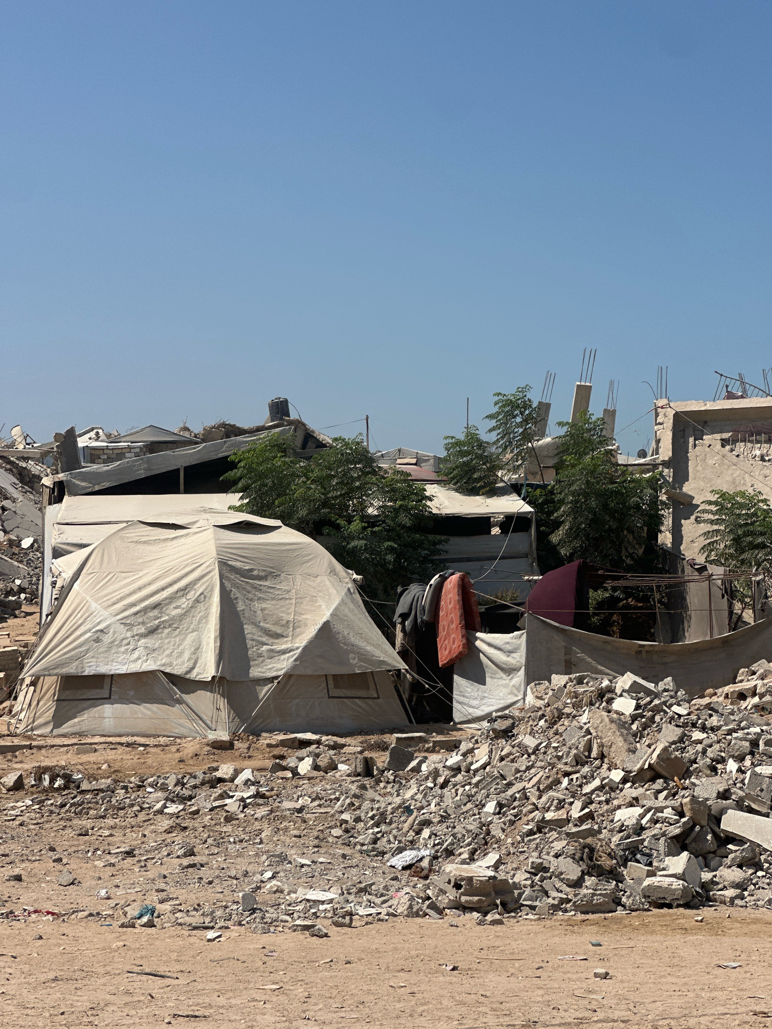 In the city of Gaza, Palestinian Territories, the image depicts a scene of destruction and makeshift shelter. In the foreground, a large, light-colored tent with multiple peaked sections stands on sandy ground. The tent has visible windows. To the right and behind the tent, a massive pile of rubble, consisting of broken concrete, rebar, and other debris, indicates the destruction of buildings. Several trees are interspersed among the remnants of structures. In the mid-ground, partially visible structures show exposed rebar and damaged walls, suggesting recent conflict or damage. Laundry, including a red towel and dark clothing, is hung on a line between structures. The sky is clear and blue, with no clouds. The environment appears arid and dusty.