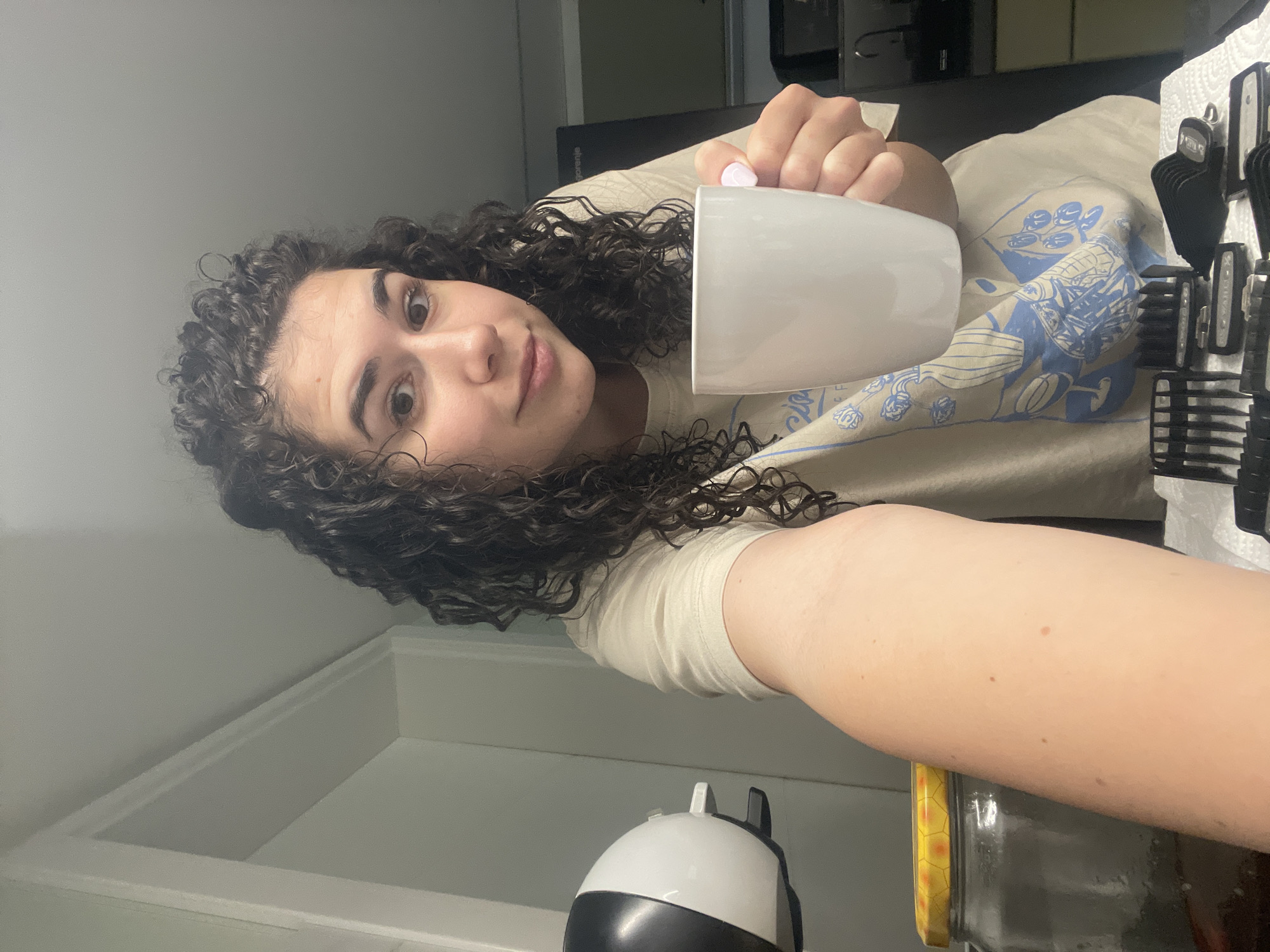 A woman with curly brown hair is taking a selfie while holding a white mug. She has fair skin and is wearing a light-colored t-shirt. In the background, there are grooming tools on a counter, a jar, and an appliance. The location is not identifiable as Sofia, Bulgaria.