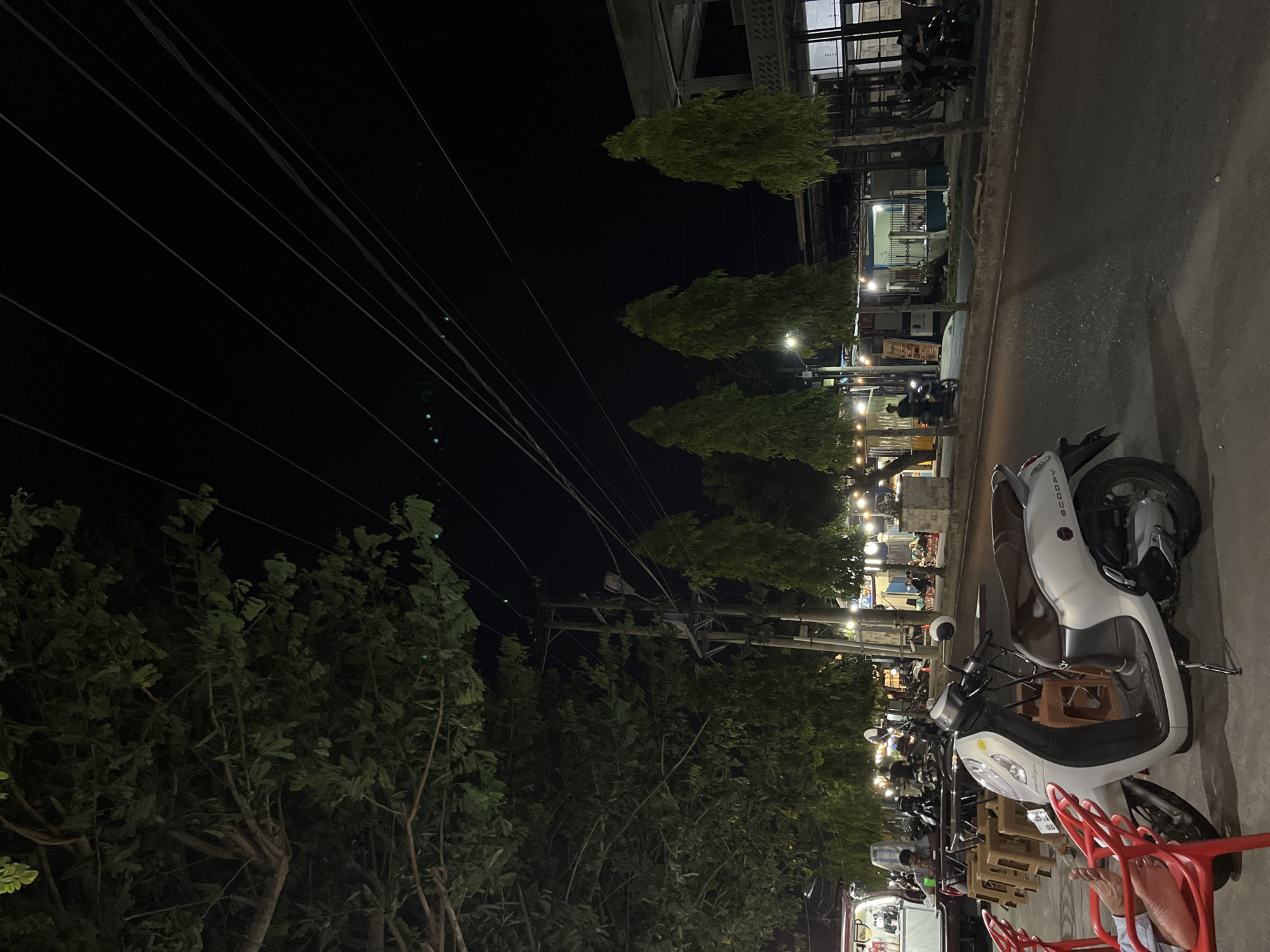 A night scene captures a street in Lhokseumawe, Indonesia, with a focus on an outdoor seating area next to a road. In the foreground, a white scooter is parked on the side of the road, with two red chairs to its left. One chair has a person's feet visible, resting on it. Behind the scooter, a row of small wooden tables and chairs are set up, with several people seated and dining. A food truck or mobile vendor is parked to the far left.
Across the street, a row of shops and food stalls are illuminated by string lights, creating a vibrant atmosphere. Tall, slender trees line the street, interspersed with utility poles and overhead power lines stretching across the dark sky. The road appears to be paved, with some parked motorcycles and a few people walking or riding. The overall impression is of a lively evening street scene in a Southeast Asian city.