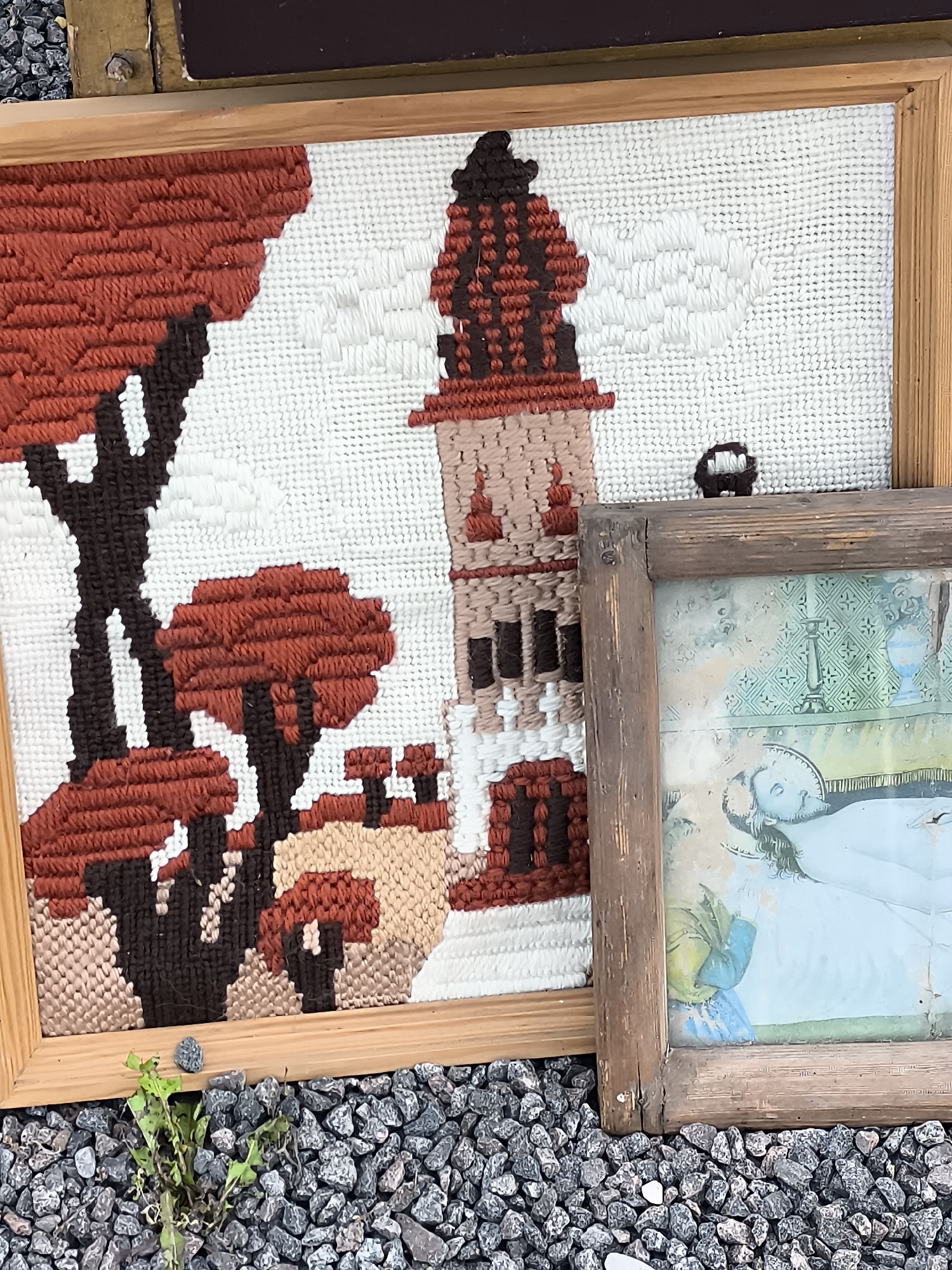 This is a close-up, eye-level, outdoor, full shot of two framed art pieces resting on a bed of gray gravel. The art on the left is a needlepoint tapestry depicting a reddish-brown tree with a dark brown trunk against a white background, with a tan and brown building and a brown tower in the distance. The frame is made of light-colored wood. To the right of the tapestry is a smaller, framed picture with a weathered, dark brown wooden frame. The artwork within this frame appears to be a faded painting or print, possibly of a person in bed, rendered in pale blue and green tones, with a candlestick and blue vase visible in the background. A small green plant is growing out of the gravel in the bottom left corner of the image. The background above the art pieces is blurred, showing what appears to be the edge of another object with a darker brown and yellow-gold color. The scene is likely from a flea market or antique shop in Klaipėda, Lithuania, given the style of the art and framing which could be indicative of local craft or historical items.