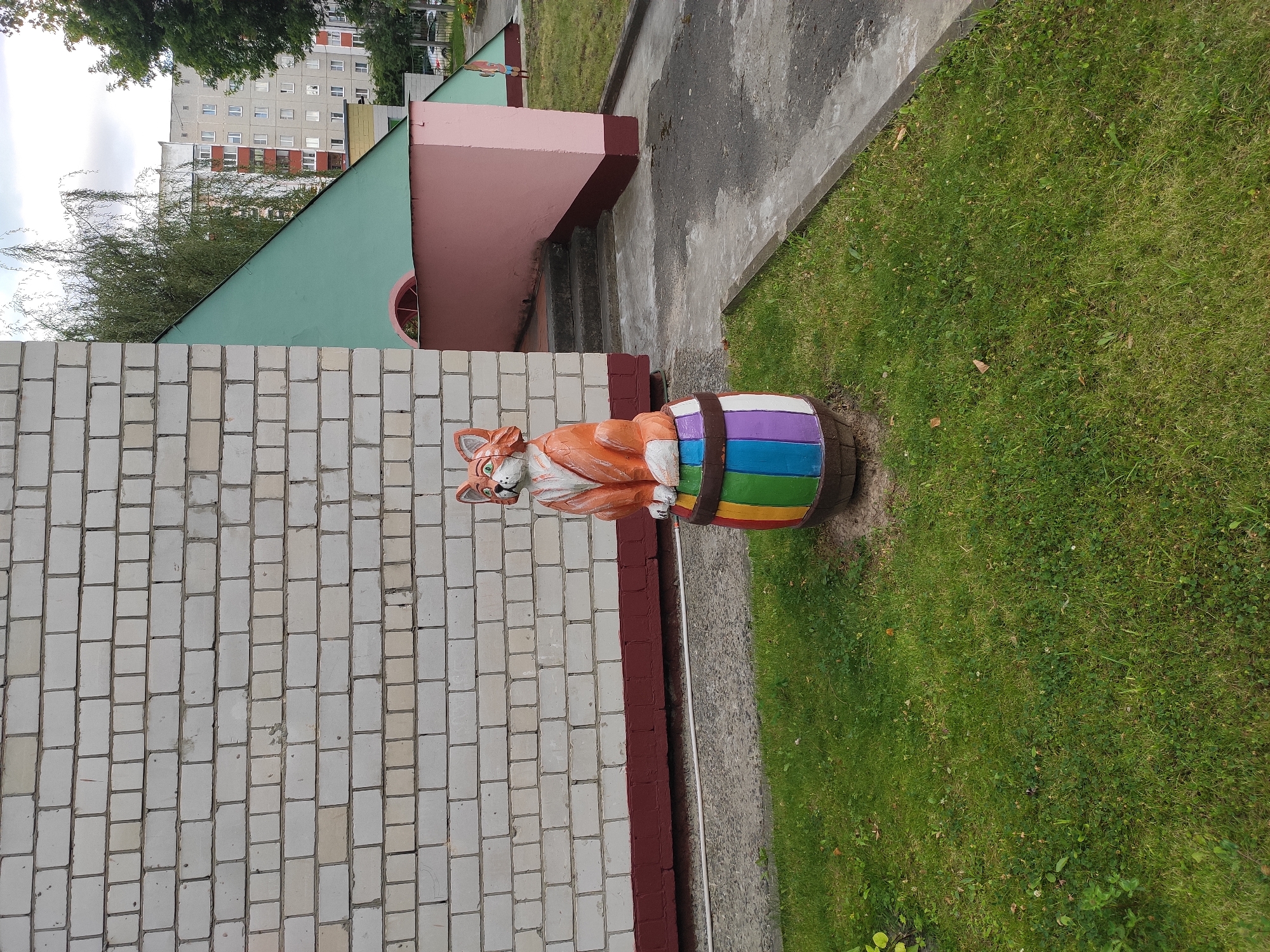 In the city of Homyel, Belarus, a whimsical fox sculpture sits atop a brightly colored barrel in front of a building with a white brick wall. The fox, rendered in orange and white, gazes upwards with green eyes. The barrel it rests on is painted with a rainbow of stripes in purple, blue, green, yellow, and white. The building's foundation is a dark red, contrasting with the lighter brickwork above. To the right of the building, a structure with a pink lower section and a green, angled roof is visible, with a set of stairs leading to an entrance. Beyond this, apartment buildings can be seen in the distance, typical of urban Belarusian architecture, under a cloudy sky. The foreground features a patch of green grass with scattered leaves and clover.