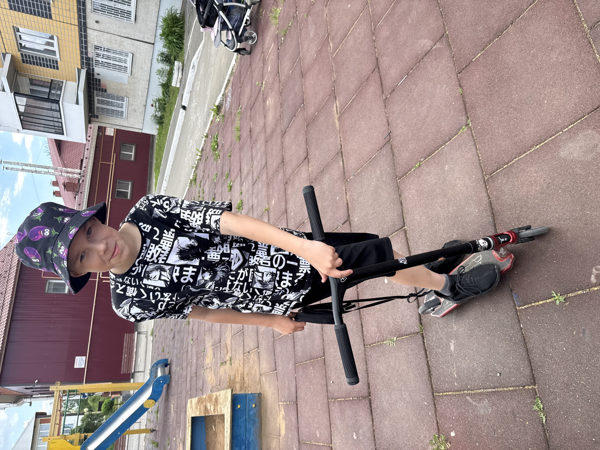 A young boy with short blonde hair, wearing a black bucket hat with an eggplant pattern and a black t-shirt with a white graphic print, stands on a red rubberized playground surface with a black scooter. He is holding the scooter's handlebars with his right hand. The boy is wearing black shorts and black sneakers with grey socks. In the background, there is a blue and yellow playground slide, a sandbox, and multi-story residential buildings, suggesting an urban or suburban environment in Zav'yalovo, Russia. A stroller is visible on the right side of the image. The sky is cloudy.