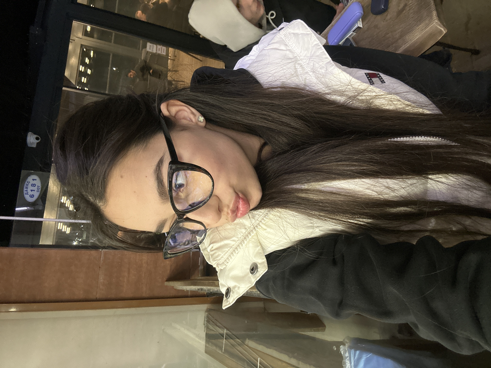 A young woman with long, dark hair is wearing glasses and posing for a selfie. She is wearing a black hoodie with a white Tommy Hilfiger jacket on top. The location is in Plovdiv, Bulgaria, as evidenced by the Cyrillic inscription "ДАРКО 6181 COT" visible in a sticker or sign behind her, likely on a window. She appears to be sitting at a table, possibly indoors by a window, as reflections suggest an outdoor environment. The table and surrounding area seem to be in a casual dining or cafe setting. Other people are visible in the background of the window reflection.