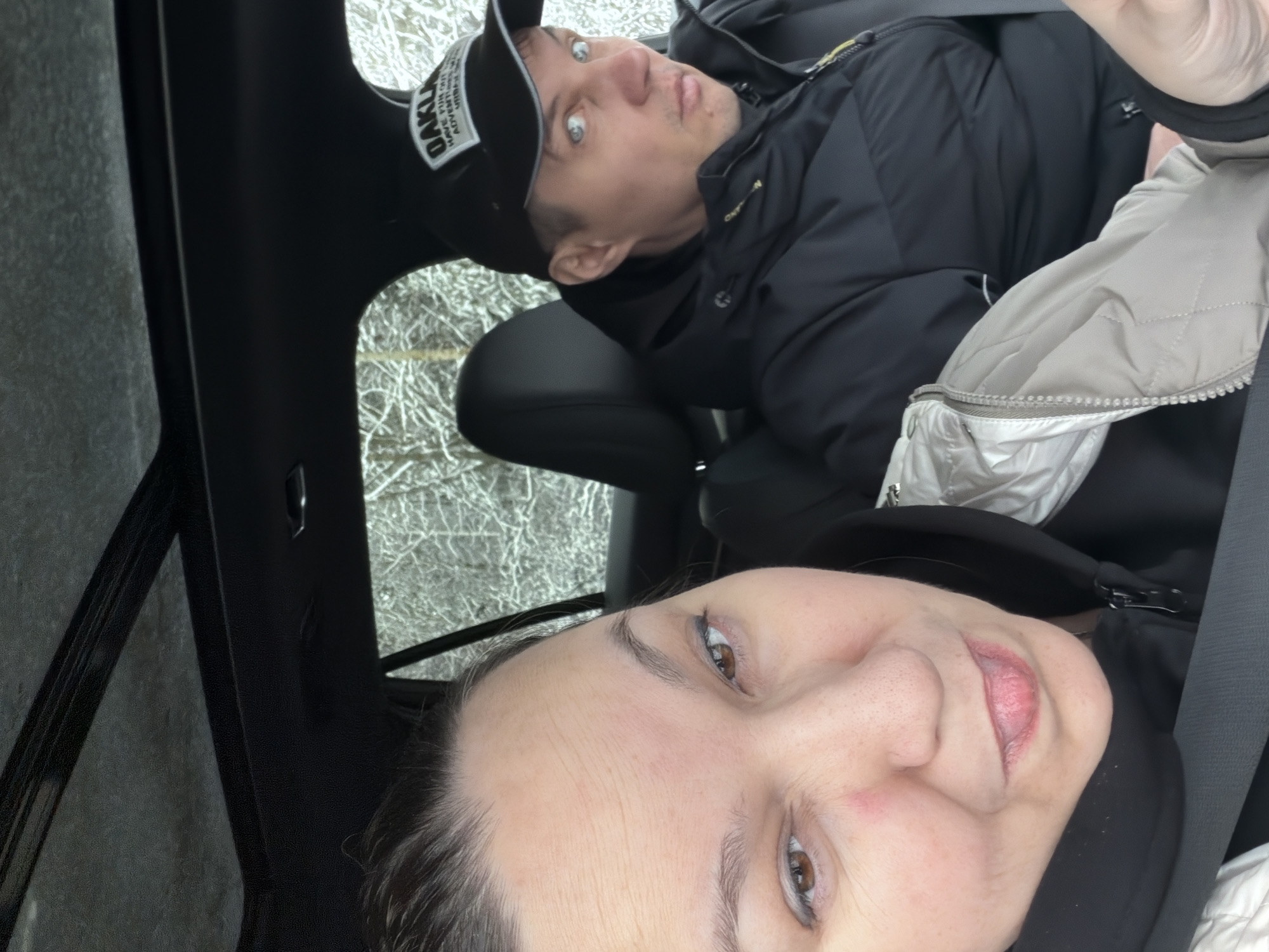 The image shows two people inside a car. A woman is in the foreground, taking a selfie. Behind her is a man wearing a black jacket and a black baseball cap with white lettering. The car's interior is black. Through the window, bare trees and branches suggest it is winter or late fall. The location is stated as Chaikovsky, Russia.