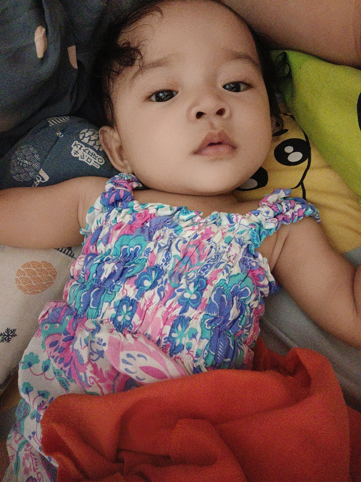 A baby lies on a bed, looking towards the camera. The baby is wearing a colorful, smocked dress. The background includes pillows and blankets. The image does not provide any information about the city of Kawua, Indonesia.