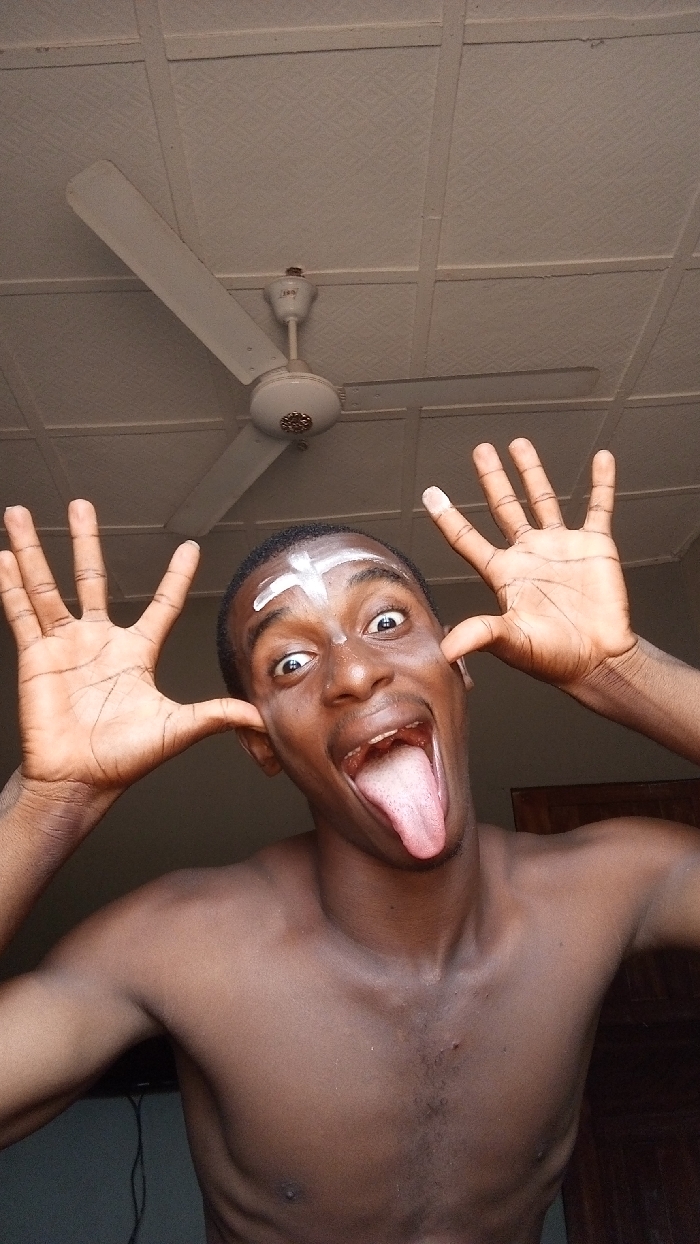 A shirtless dark-skinned man with a white cross painted on his forehead makes a silly face. He pulls his ears, widens his eyes, and sticks out his tongue. A ceiling fan is visible in the background, along with a textured ceiling and parts of a room.