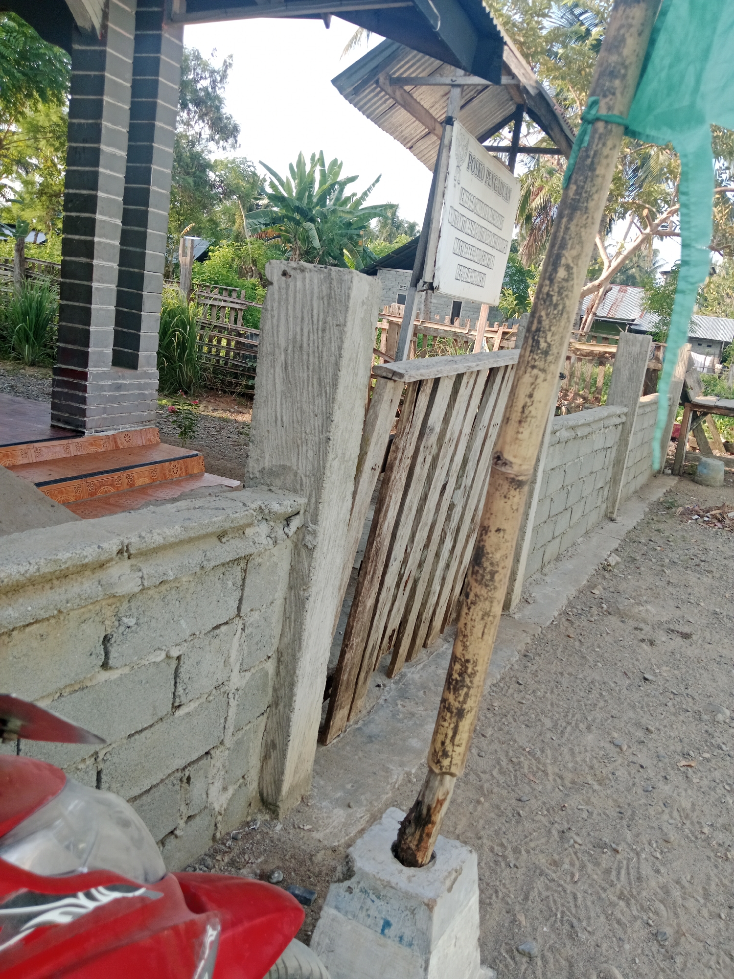 The image captures an outdoor scene, likely in Lakea Satu, Indonesia, with a prominent red motorcycle in the foreground. To the left, a building structure is visible, featuring a pillar with a black and gray checkered pattern and tiled steps. Adjacent to this, a low wall constructed from grey concrete blocks runs horizontally across the frame.
Behind the wall, a weathered wooden fence or gate is partially open, leaning against a rough concrete support. Further into the background, a variety of lush green vegetation, including what appear to be banana plants, is present. Above the vegetation, the roof of a building with corrugated metal sheeting is visible.
A sign with Indonesian text, possibly indicating an information post or station, is mounted on a wooden frame under a simple shelter. A bamboo pole, weathered and stained, stands vertically in the center-right of the image, secured in a concrete base. A piece of light green fabric or netting is tied to this pole and extends upwards and to the right. The ground is a mixture of dirt and small stones. The overall impression is a rustic, possibly rural, setting under daylight conditions.
