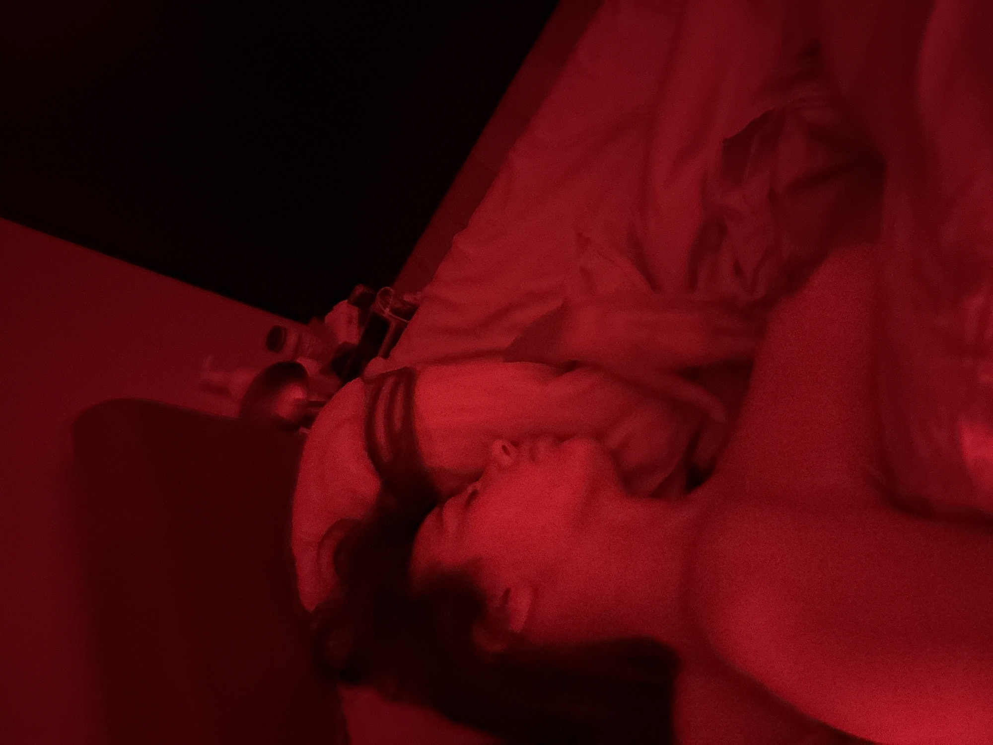 A person is lying in bed. The room is lit by a red light. There are various objects on a surface beside the bed. The location is Unknown, Unknown.