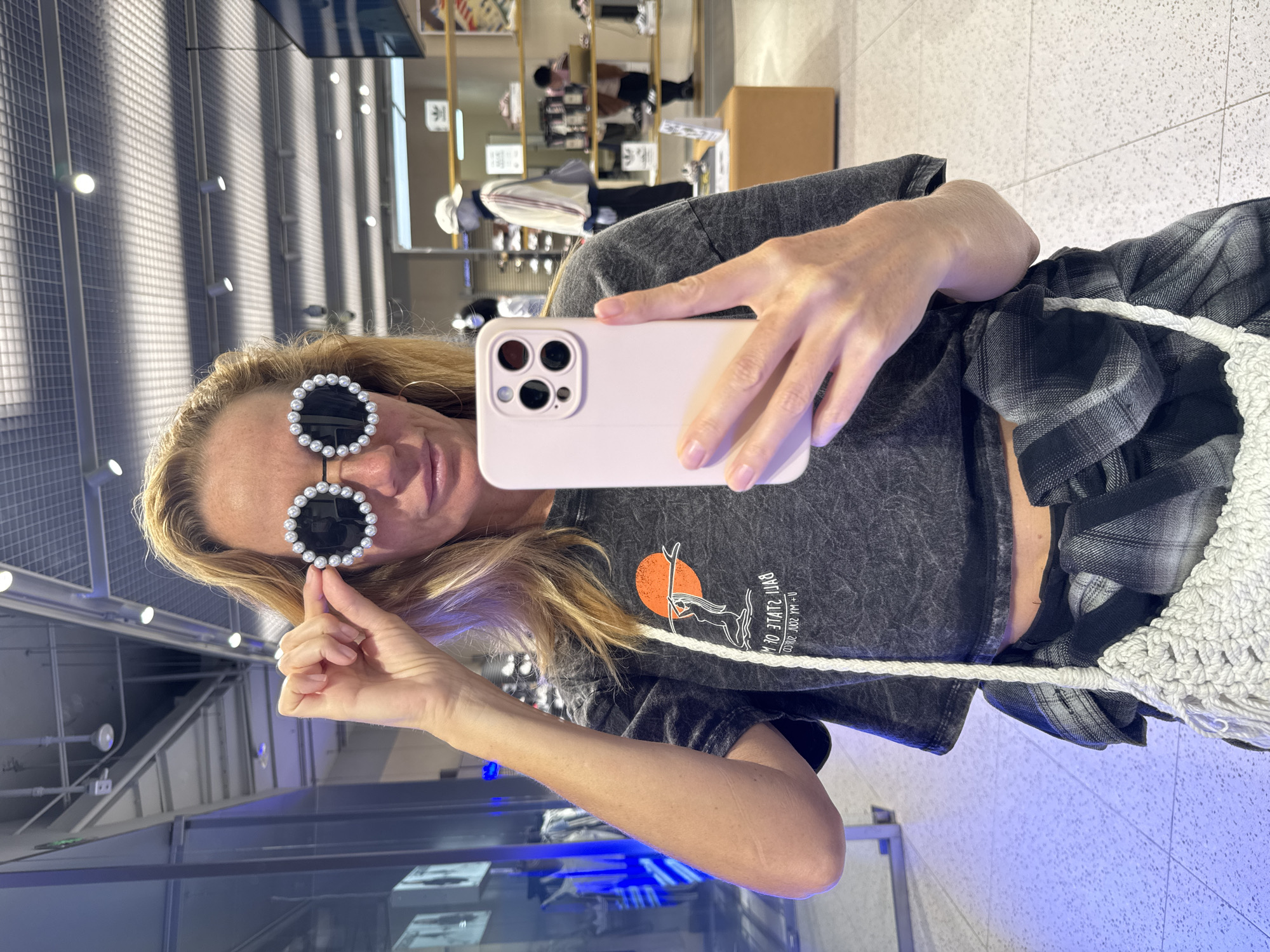 A woman takes a selfie indoors, possibly in a retail environment. She is holding a light pink smartphone with three camera lenses visible on the back. She is wearing sunglasses with pearl embellishments and a gray shirt with a graphic on the front. Part of a plaid shirt is also visible, along with a white macrame bag strap. The background shows a retail setting with shelves displaying merchandise. The location may be in Jinjiang District, China, based on potential retail settings in that area.