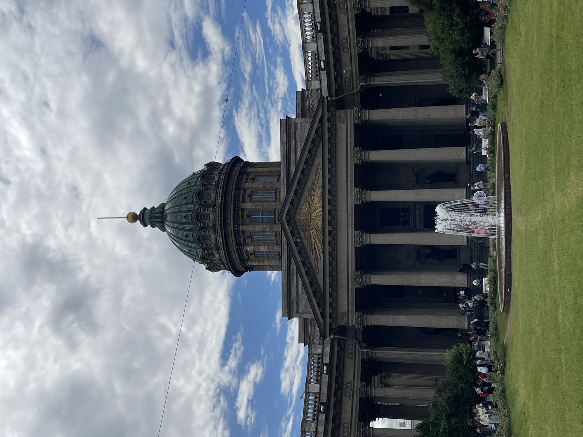 The image depicts a view of the Kazan Cathedral in Saint Petersburg, Russia. A large, green dome is visible on the left side of the frame, topped with a spire. The cathedral's facade, characterized by a semi-circular colonnade and classical architecture, extends across the right side of the image. A fountain is in the foreground, with water spraying upwards. Green grass covers the area in front of the building. Several people are seated on benches and along the edge of the grass, some facing the cathedral, others looking towards the fountain. The sky is partly cloudy with white clouds scattered against a blue background. The lighting suggests it is daytime, likely afternoon. The scene conveys a sense of people enjoying a pleasant day outdoors in a historic urban setting.