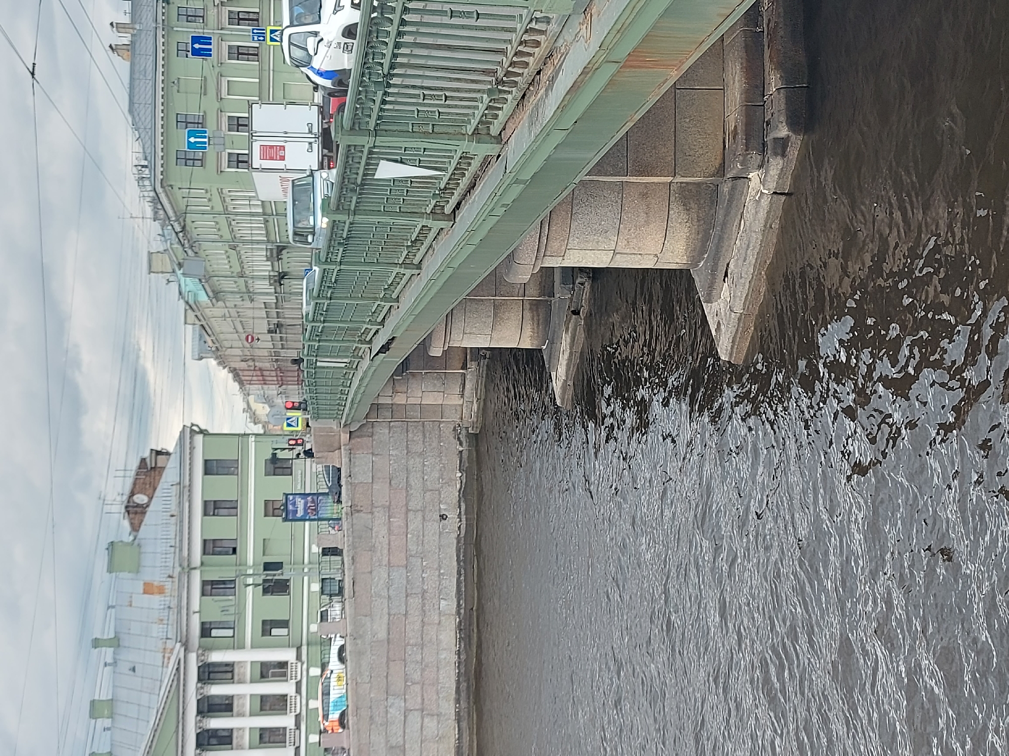 The image depicts a view of a bridge over a canal in Saint Petersburg, Russia. The bridge has a green metal railing and supports made of stone blocks that go down into the water. Cars, including what appears to be a police vehicle, are visible on the bridge. On the left, there are buildings with a light green facade and multiple windows. The water in the canal is dark and has ripples, indicating movement. The sky is overcast.