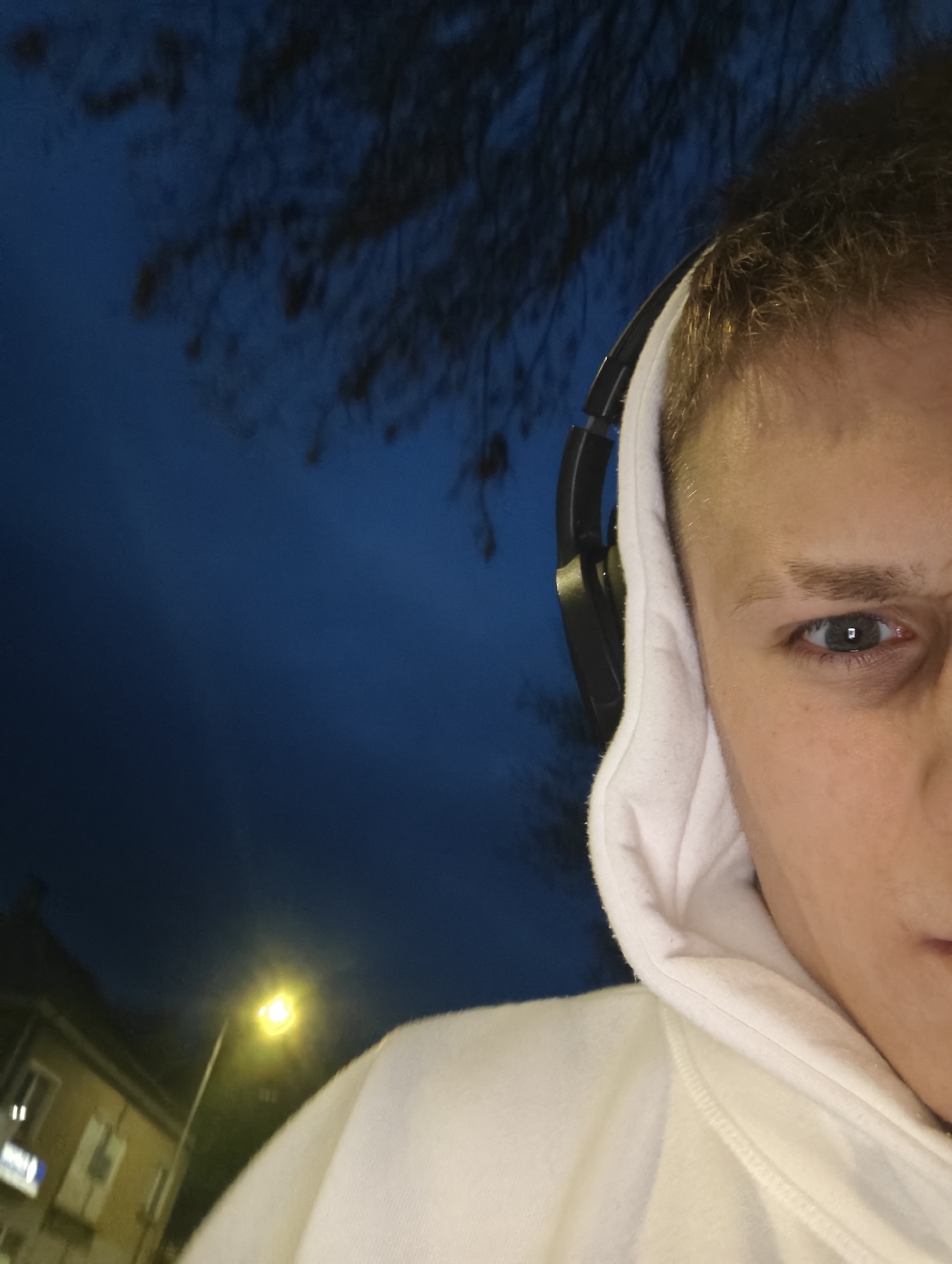 A selfie, cropped from the right, of a light-skinned person wearing a white hooded sweatshirt and black headphones is framed against a dark blue sky with tree branches visible in the upper portion. A building with a light pole near it is visible in the lower left of the frame, suggesting a nighttime setting. The image is possibly taken near Kozloduy, Bulgaria, as requested.