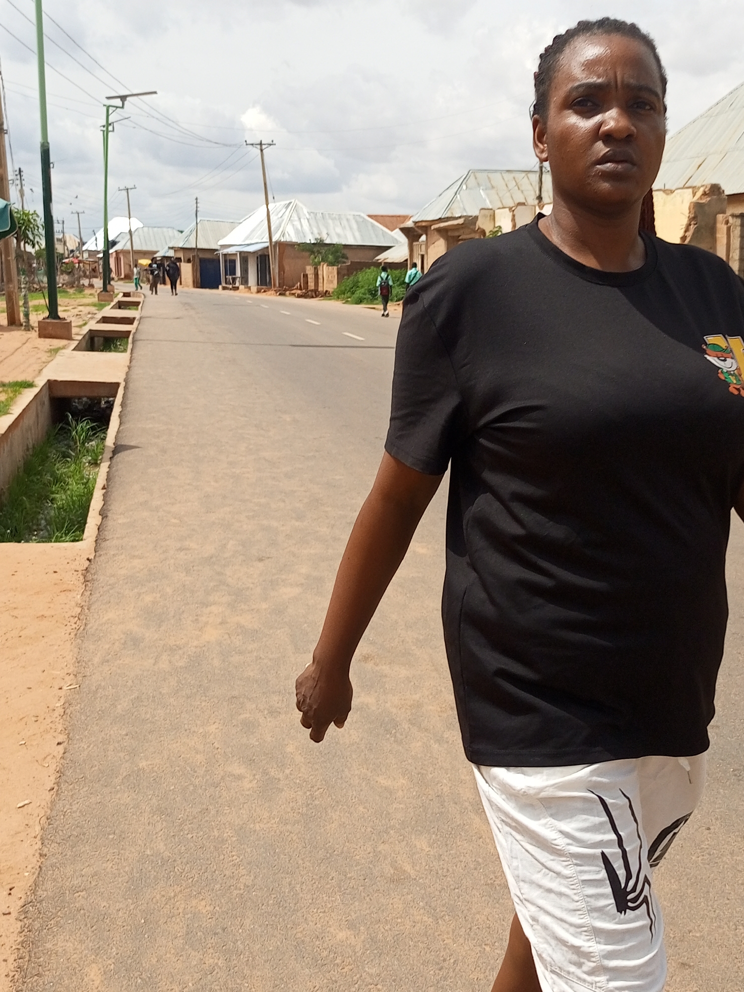 A medium shot captures a woman with dark skin and braided hair walking on an asphalt road. She is wearing a black t-shirt with a cartoon graphic on the right chest and white shorts with a black spider design. The woman is looking forward with a serious expression. The background shows a row of buildings with corrugated metal roofs and some vegetation along the roadside. Power lines and streetlights are visible overhead. The scene appears to be in Gombe, Nigeria, based on the architectural style and infrastructure. The road has white markings and a drainage ditch runs along the left side. There are a few other people walking in the distance.