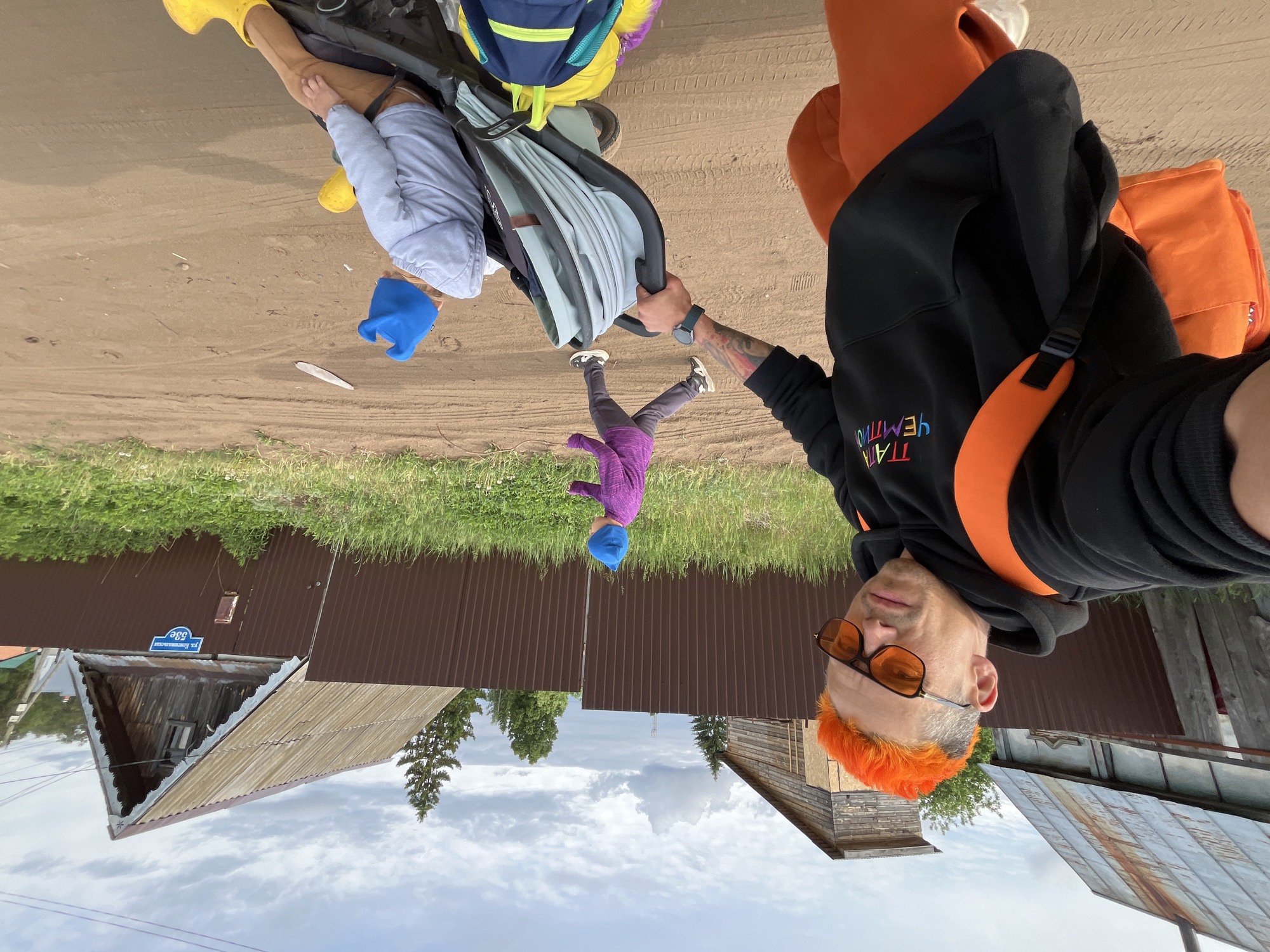 A photograph taken in Chaikovsky, Russia, presents a family walking along a dirt road adjacent to a fence. The perspective is unusual, as the camera appears to be held upside down. The foreground features an adult wearing a black hoodie with colorful lettering, orange pants, and orange hair, looking at the camera. The adult is holding a stroller. Inside the stroller is a child wearing a blue hat and tan Crocs. A young child wearing a purple outfit and blue hat is walking in front of the stroller. A large orange backpack is draped over the stroller. The background includes a brown metal fence, grassy areas, wooden buildings, and a cloudy sky. A blue street sign is visible on one of the buildings. The dirt road is slightly uneven and shows traces of vehicle tire tracks.
