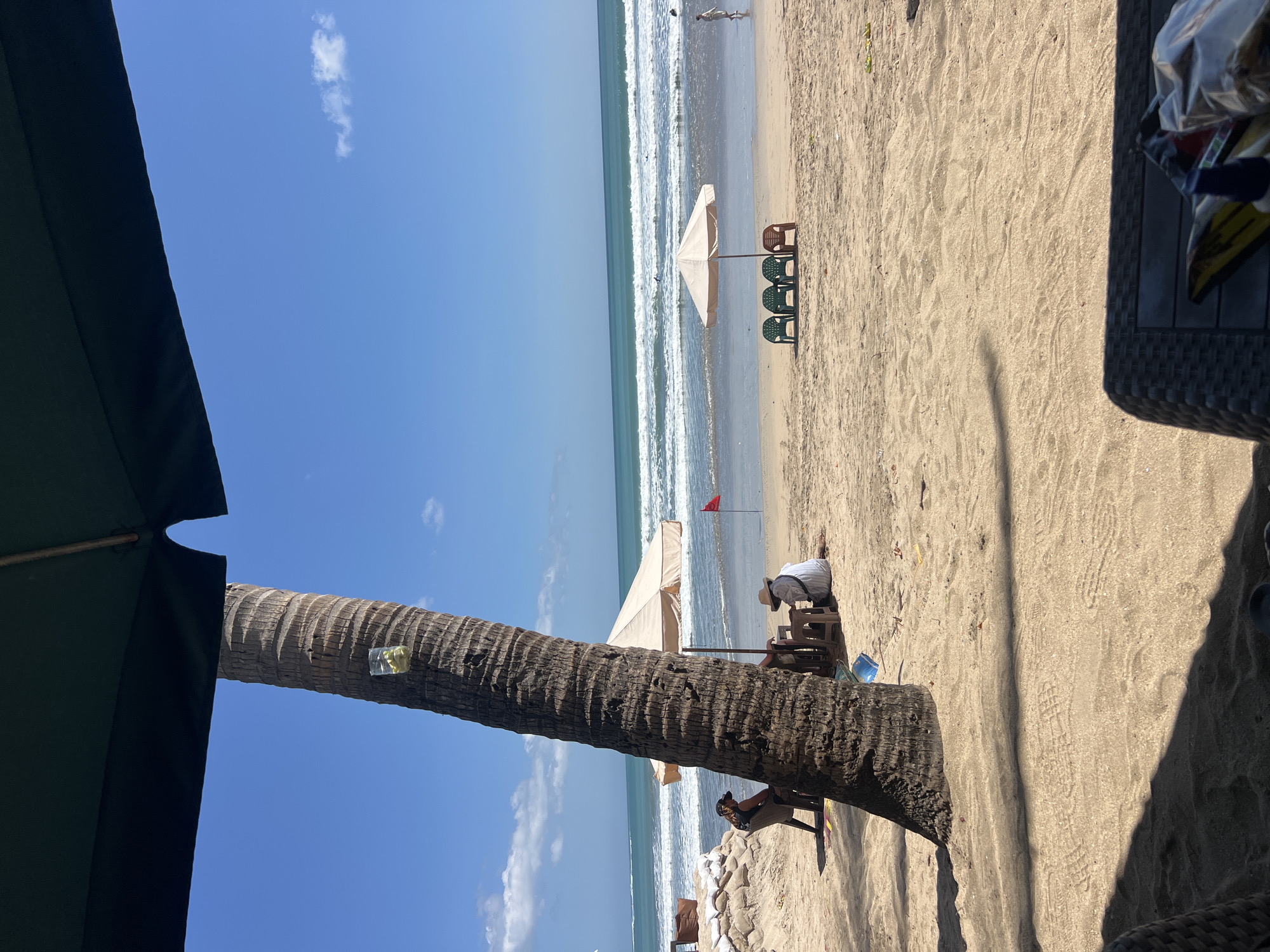 This image depicts a tropical beach scene in Kuta, Indonesia, under a clear blue sky with sparse clouds. In the foreground, a large palm tree stands prominently on the sandy beach, with a clear plastic cup containing a light-colored liquid and green fruit slices attached to its trunk. Partially visible in the foreground is the edge of a dark-colored woven chair and a table.
Further back, on the beach, two individuals are seated at a table under a beige umbrella. One person is wearing a hat and a white top, while the other appears to be wearing a black top and a dark baseball cap. A red flag is planted in the sand behind them.
In the mid-ground, another beige umbrella is set up, accompanied by three green plastic chairs and one brown plastic chair. The ocean stretches out to the horizon, with gentle waves rolling onto the shore. Several surfers can be seen in the water. On the right side of the frame, a person is walking along the shoreline.
The background consists of the ocean and the sky, with a few white clouds scattered across the blue expanse. The overall atmosphere is one of a relaxed beach day.
