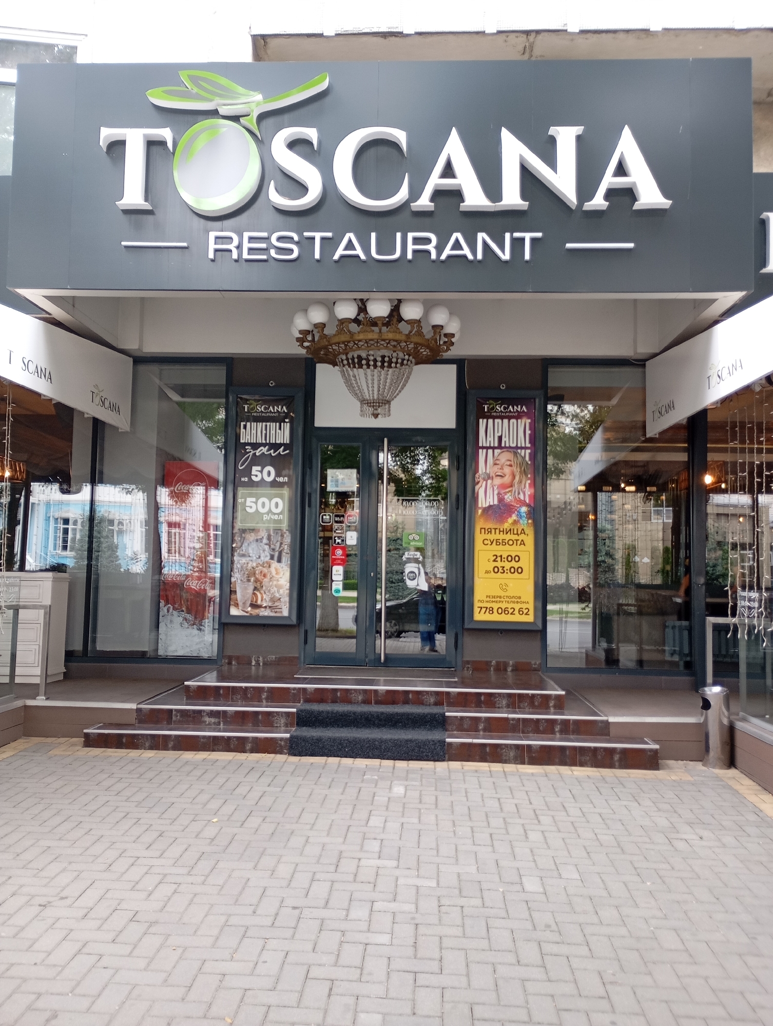 The image shows the exterior of a restaurant named "TOSCANA RESTAURANT". The restaurant has a prominent sign with the name in large white letters and a green dragonfly logo above it. Below the main sign, "RESTAURANT" is also written in white letters.
The entrance to the restaurant features a set of stairs leading up to a glass-fronted building. On the left side of the entrance, there are large windows, one of which displays a Coca-Cola advertisement. To the right of the entrance, two vertical posters are displayed. The first poster advertises a "BANQUET HALL" for 50 people at 500 rubles per person. The second poster promotes "Karaoke" on Fridays and Saturdays from 21:00 to 03:00, with a phone number for table reservations.
The overall scene depicts the daytime exterior of a commercial establishment, likely in a city. The lighting suggests it is daytime, and the presence of awnings and clear skies implies fair weather. The architecture and signage are modern. The cobblestone pavement in front of the restaurant and the visible trash can provide additional details about the immediate surroundings. No people are clearly visible in the main part of the image, though a person's legs can be seen through the glass door on the far left.