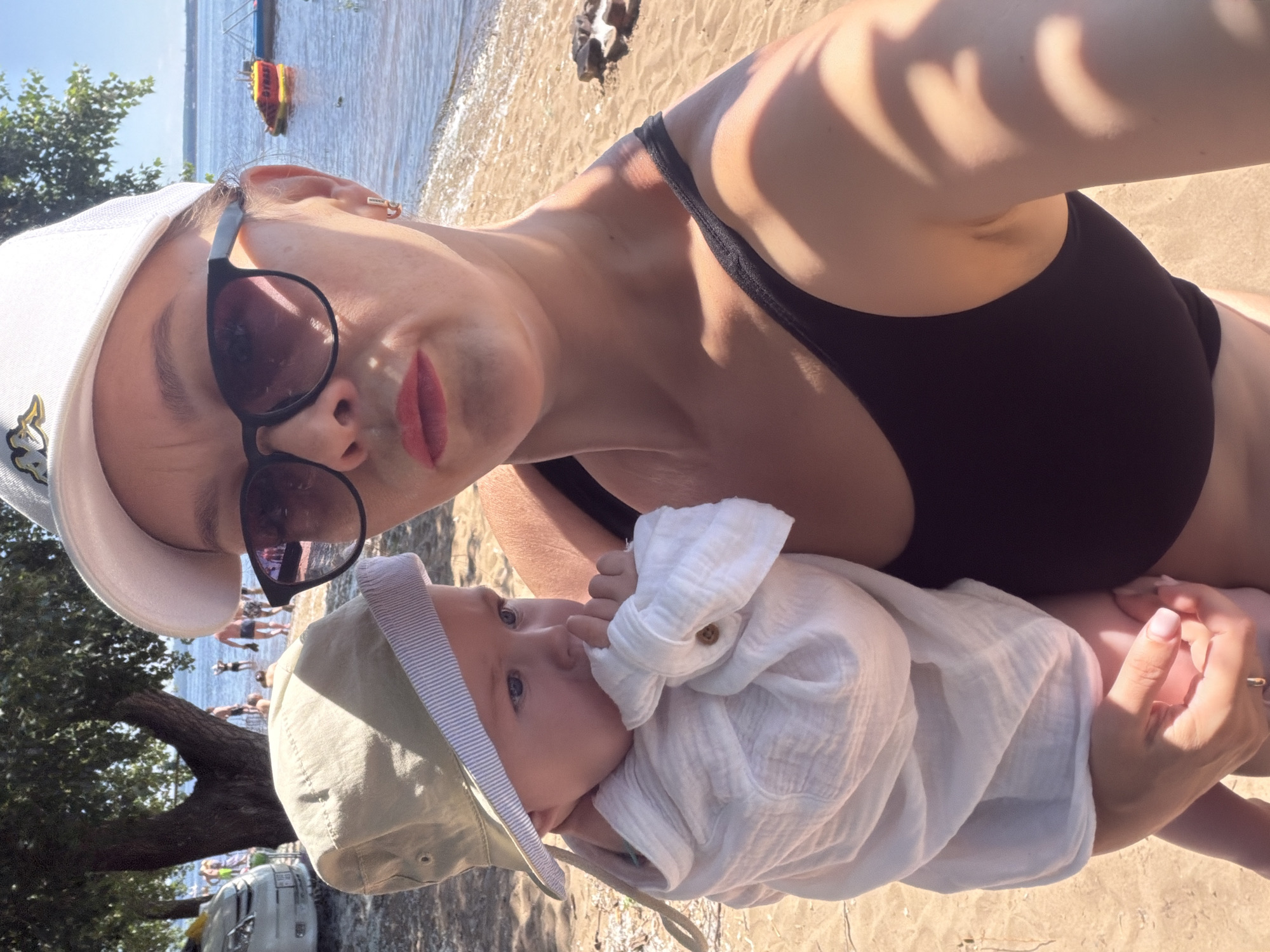A woman and a baby are on a beach in Noviy, Russia. The woman is wearing a black swimsuit, sunglasses, and a white baseball cap. She is holding a baby, who is wearing a light-colored hat and a white shirt. The baby is looking away from the camera and has its hand near its mouth. In the background, there are trees, people swimming and wading in the water, and a boat. The beach is sandy, with some small rocks and debris. The water is calm and reflects the sunlight. The sky is clear and blue.