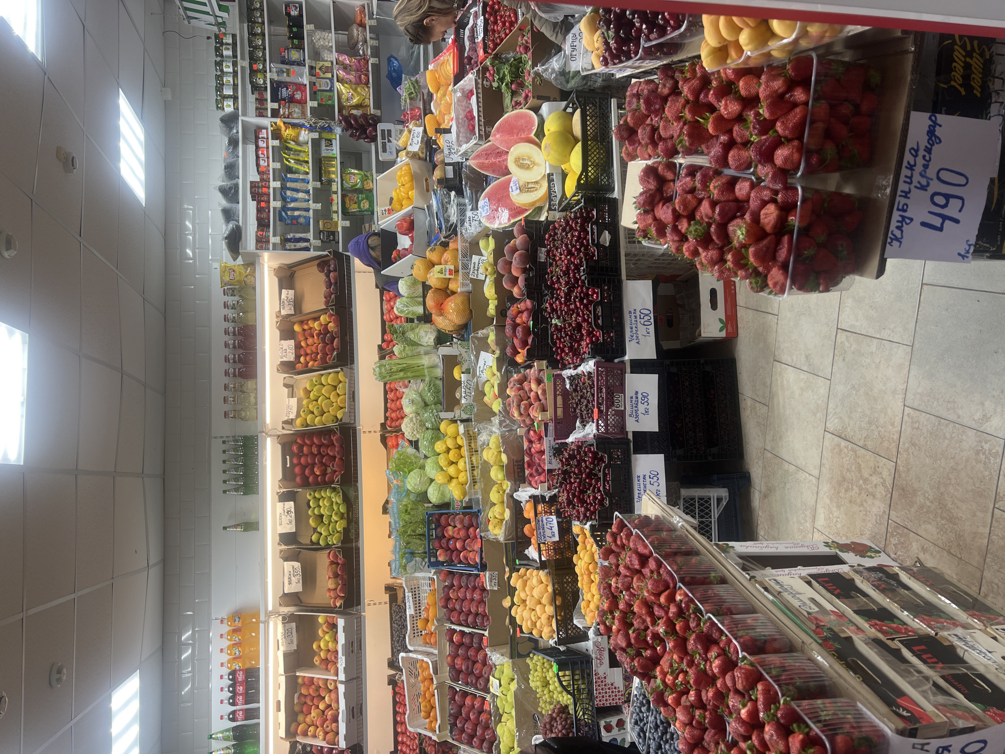The image shows the interior of a fruit market in Moscow, Russia. The market is well-stocked with a wide variety of fruits, including apples, peaches, plums, grapes, cherries, and strawberries. The produce is arranged neatly in crates and on shelves. Price tags are prominently displayed, indicating the cost of the fruits per kilogram. A person with light-colored hair, presumably a customer, can be seen in the background near a shelf stocked with packaged goods. The market appears to be well-lit and clean, with tiled floors and walls. The overall impression is one of a vibrant and organized retail space.